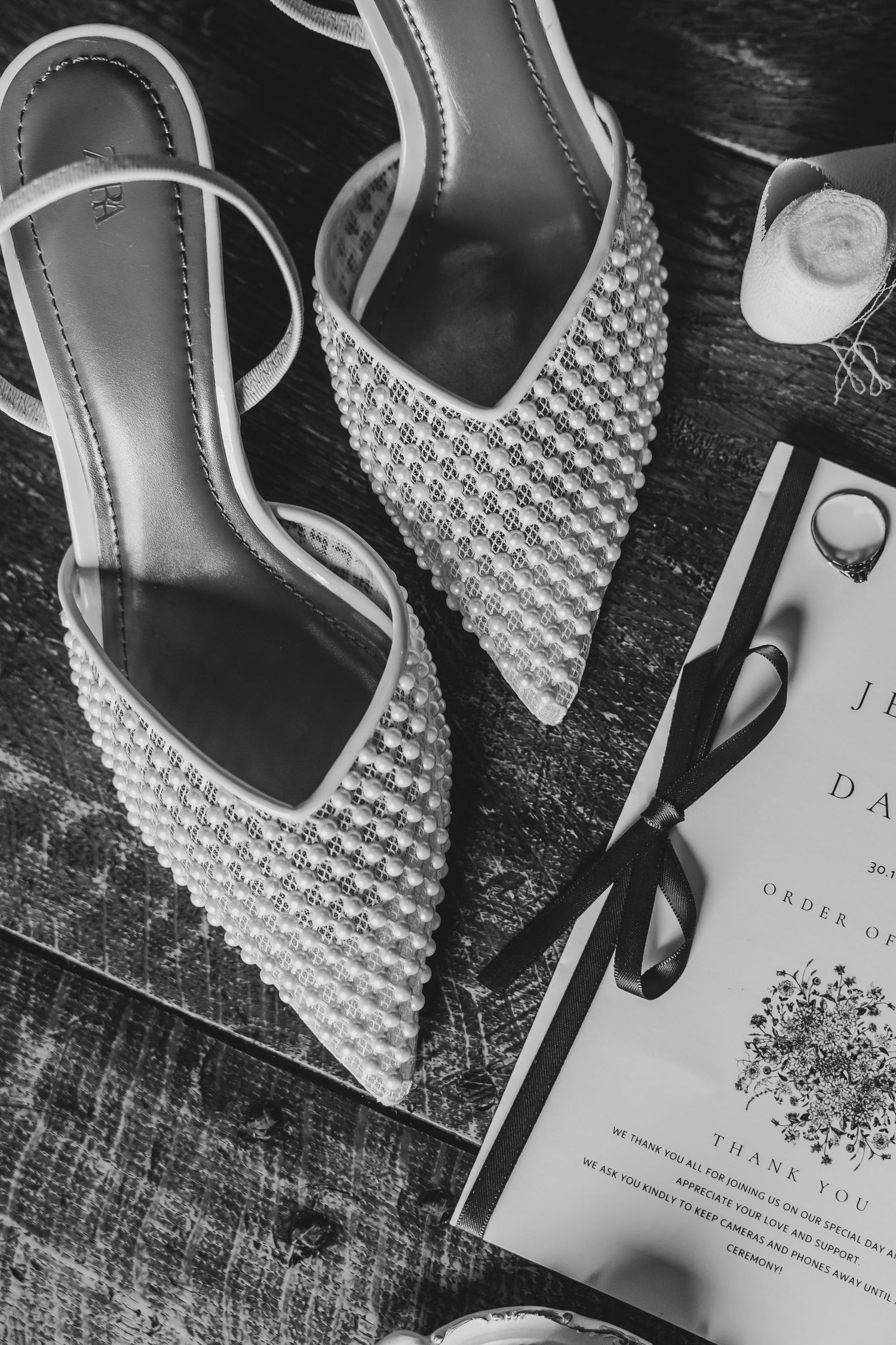 Black and white photo of pearly bridal shoes and wedding invitation on wooden background.