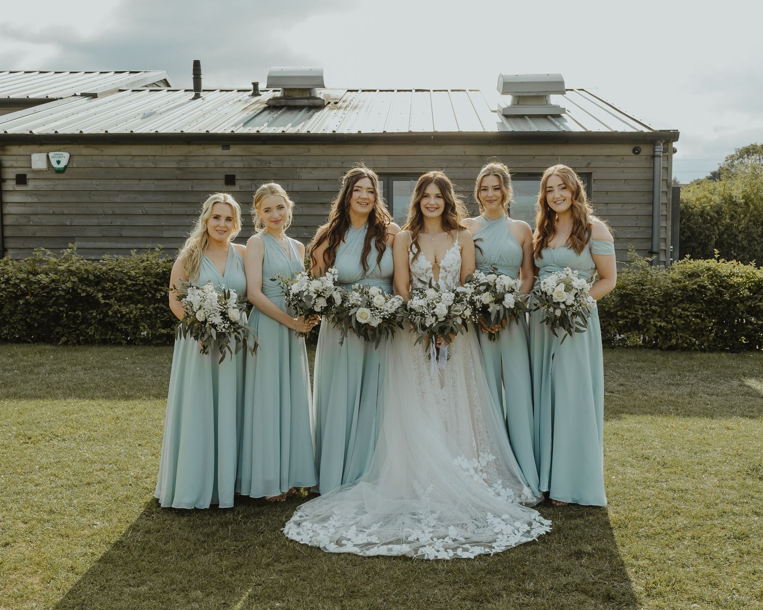 Bridesmaids in light blue dresses stand with bride in white lace gown holding floral bouquets.