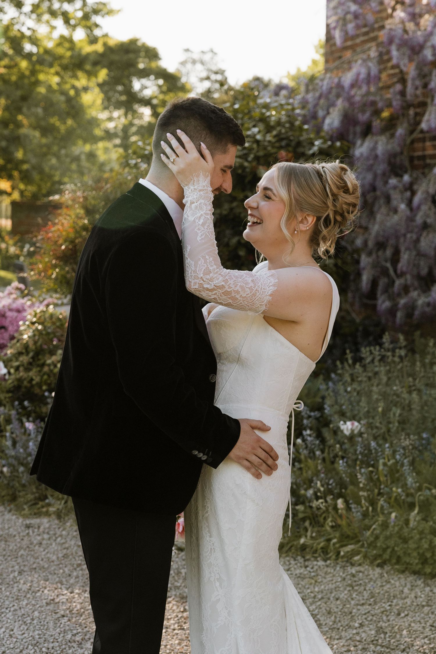 Bride in white lace gown and groom in black suit share romantic moment in garden with wisteria blooms.