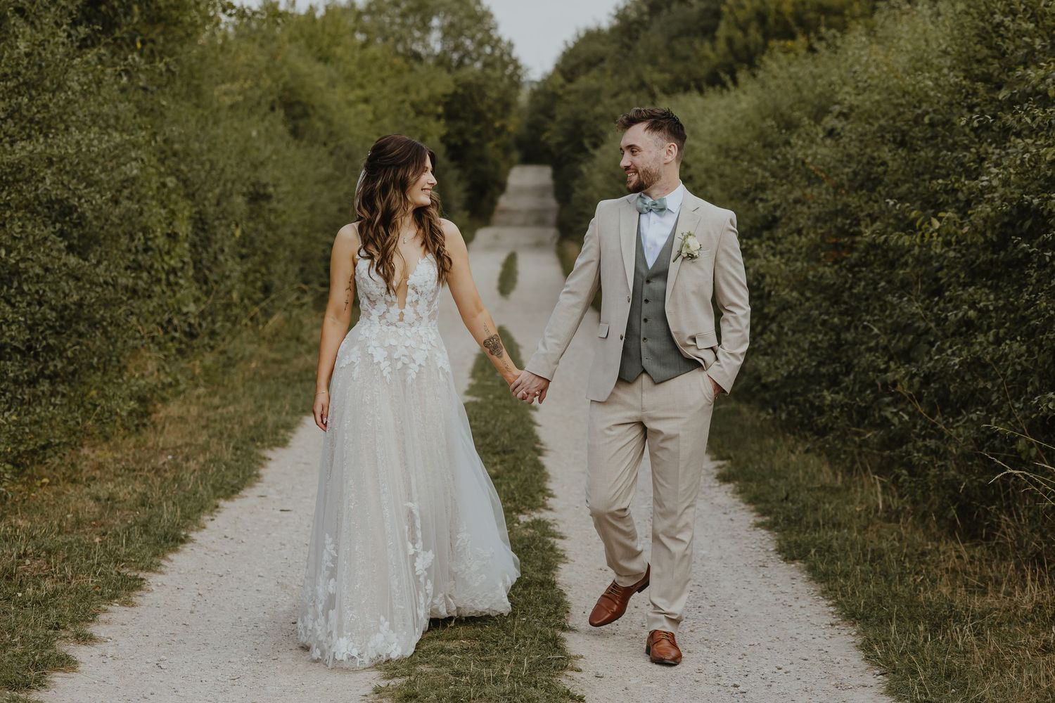 Newlyweds walk together on a sunlit path surrounded by summer foliage.