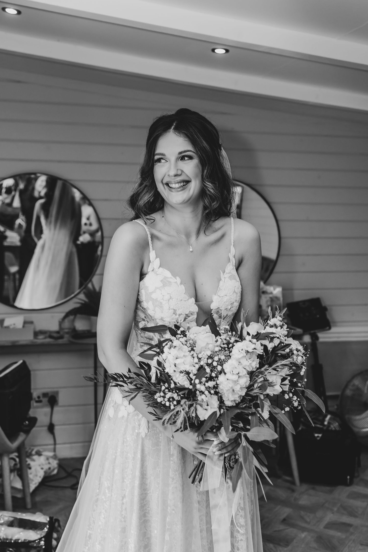 A bride wearing a white lace gown holds her bouquet while smiling in a getting ready room.