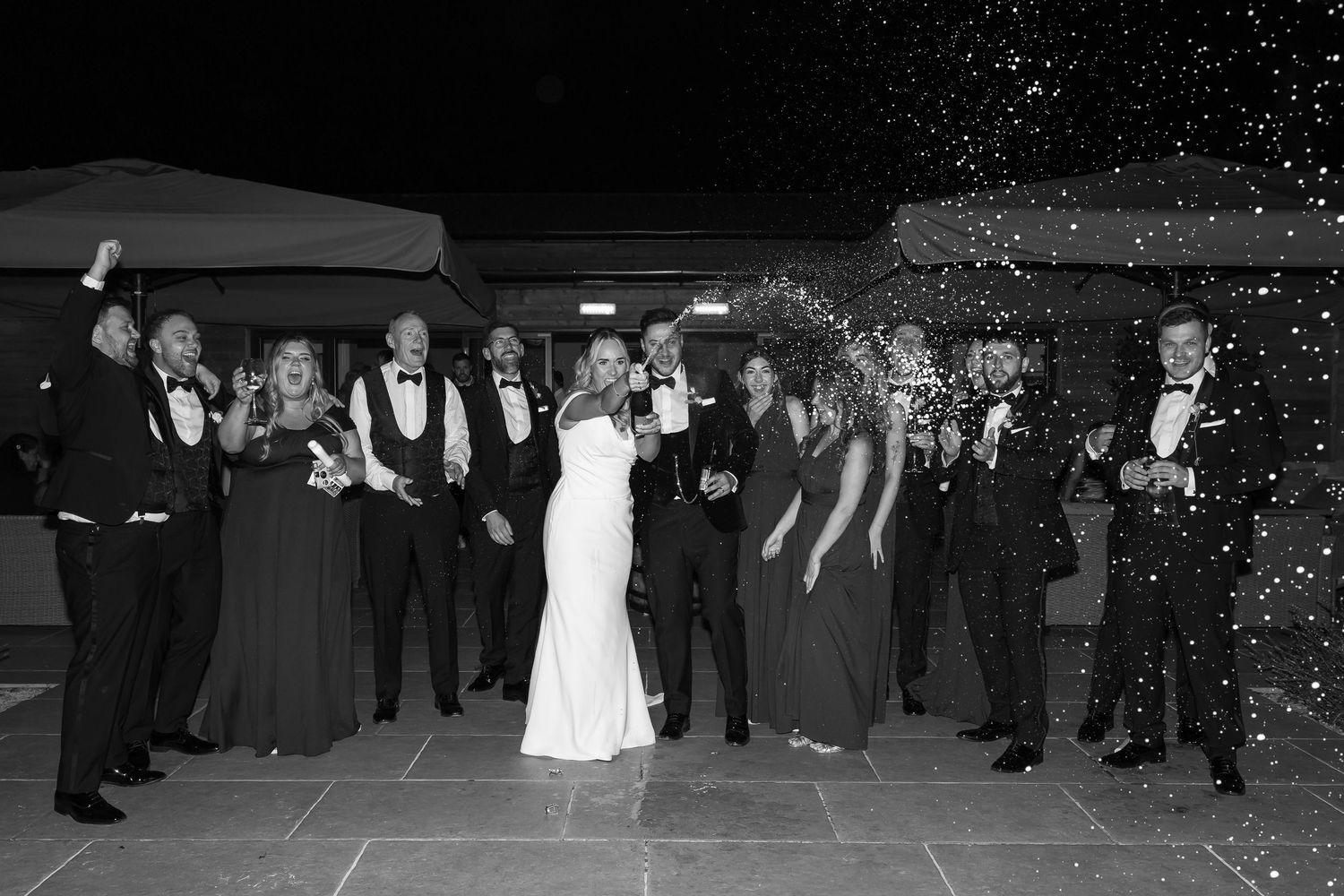 Black and white wedding photo of bride, groom, and wedding party celebrating outdoors with champagne spray.