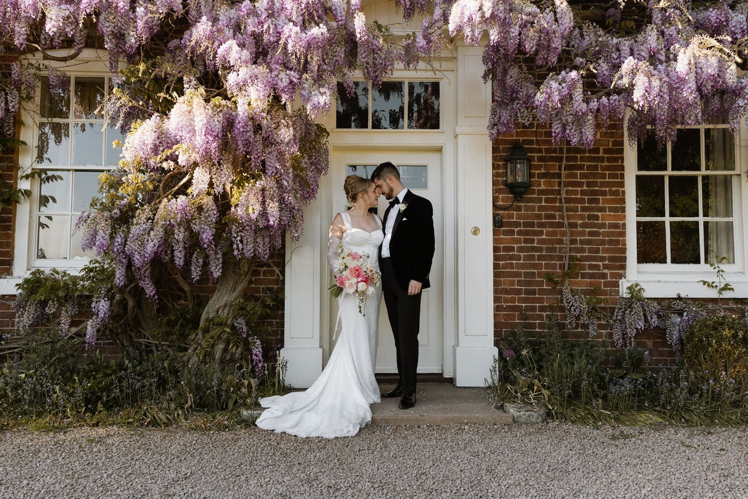 Bride and groom share a romantic moment under blooming purple wisteria at a brick manor house wedding venue.