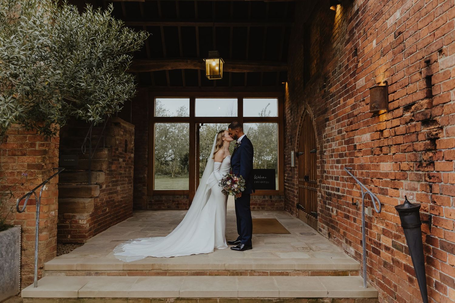 Couple shares intimate moment against rustic brick wall in wedding attire at historic venue.