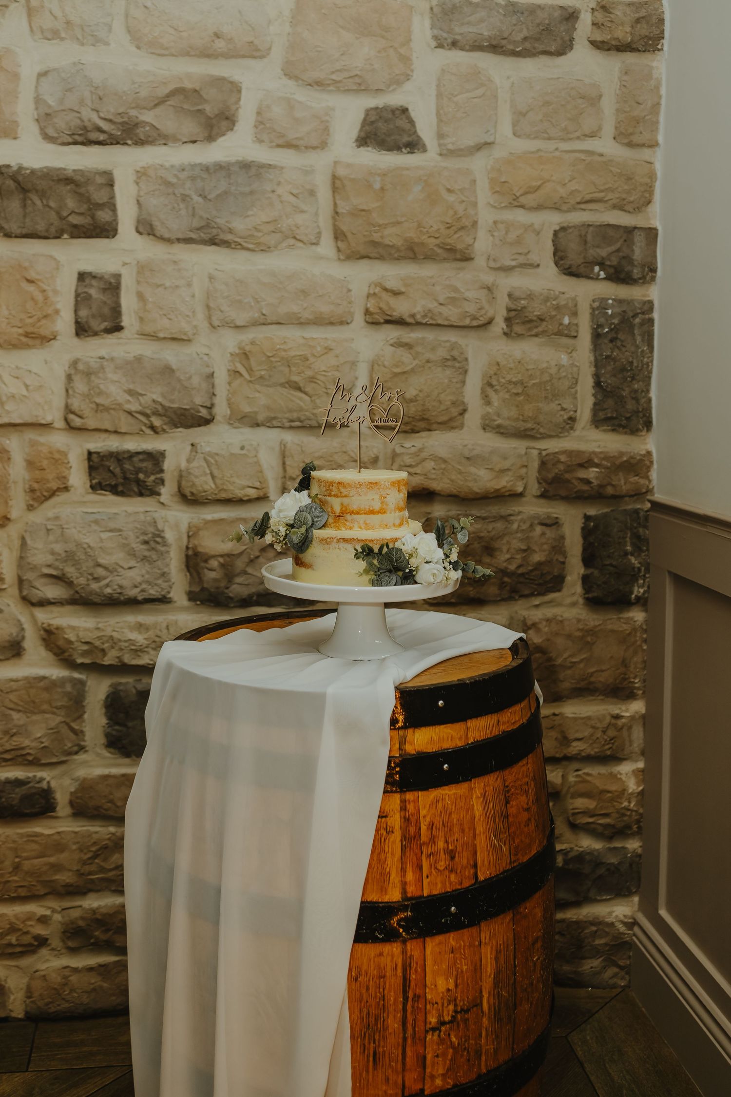 A simple two-tier white wedding cake displayed on a wooden barrel against a stone wall.