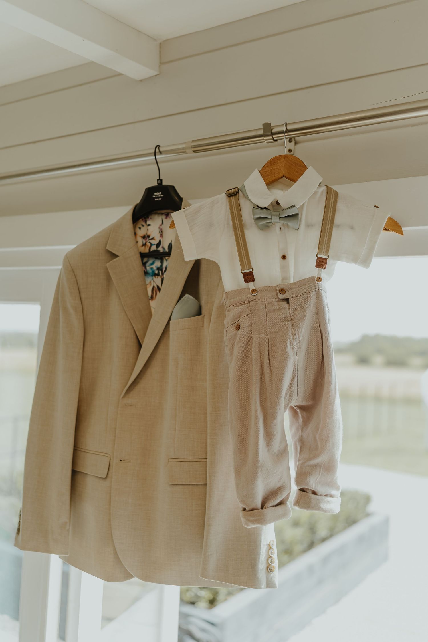 Beige and grey formal suits hanging on wooden hangers against a white wall.