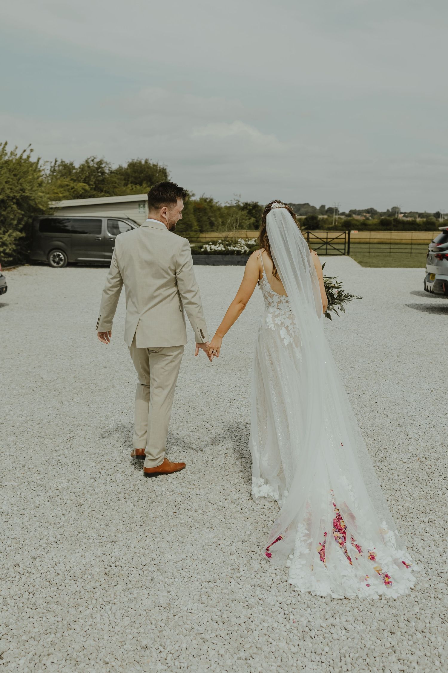 Couple walks hand in hand across white gravel path with flowing dress trailing behind.