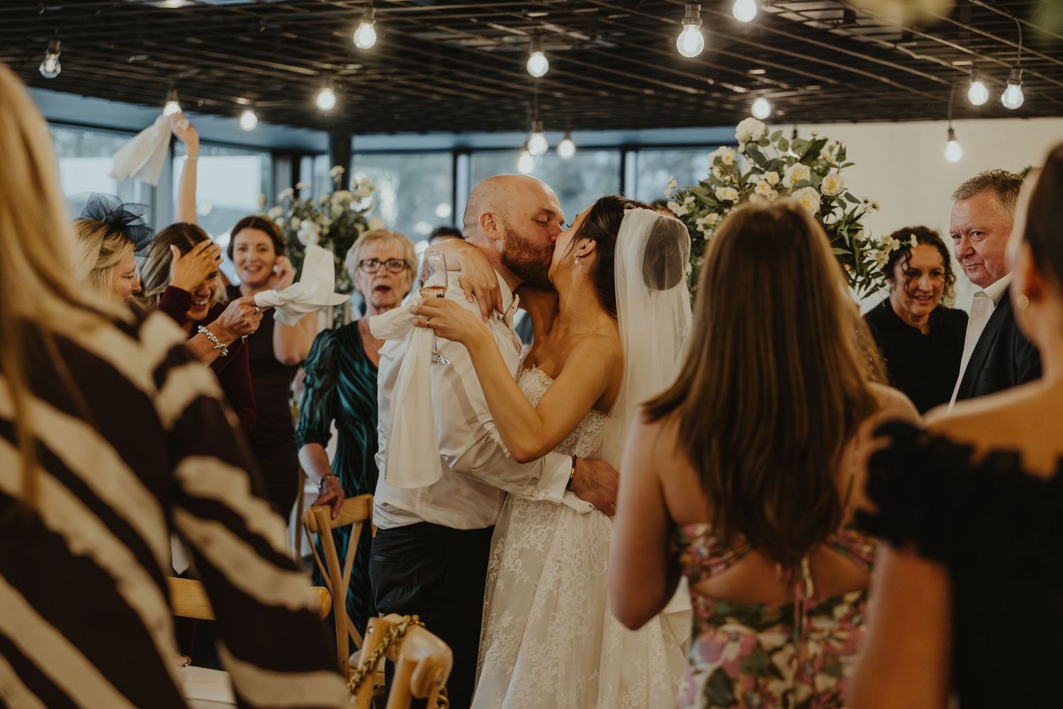 Wedding reception guests dance and celebrate on a dimly lit dance floor under string lights at an evening party.