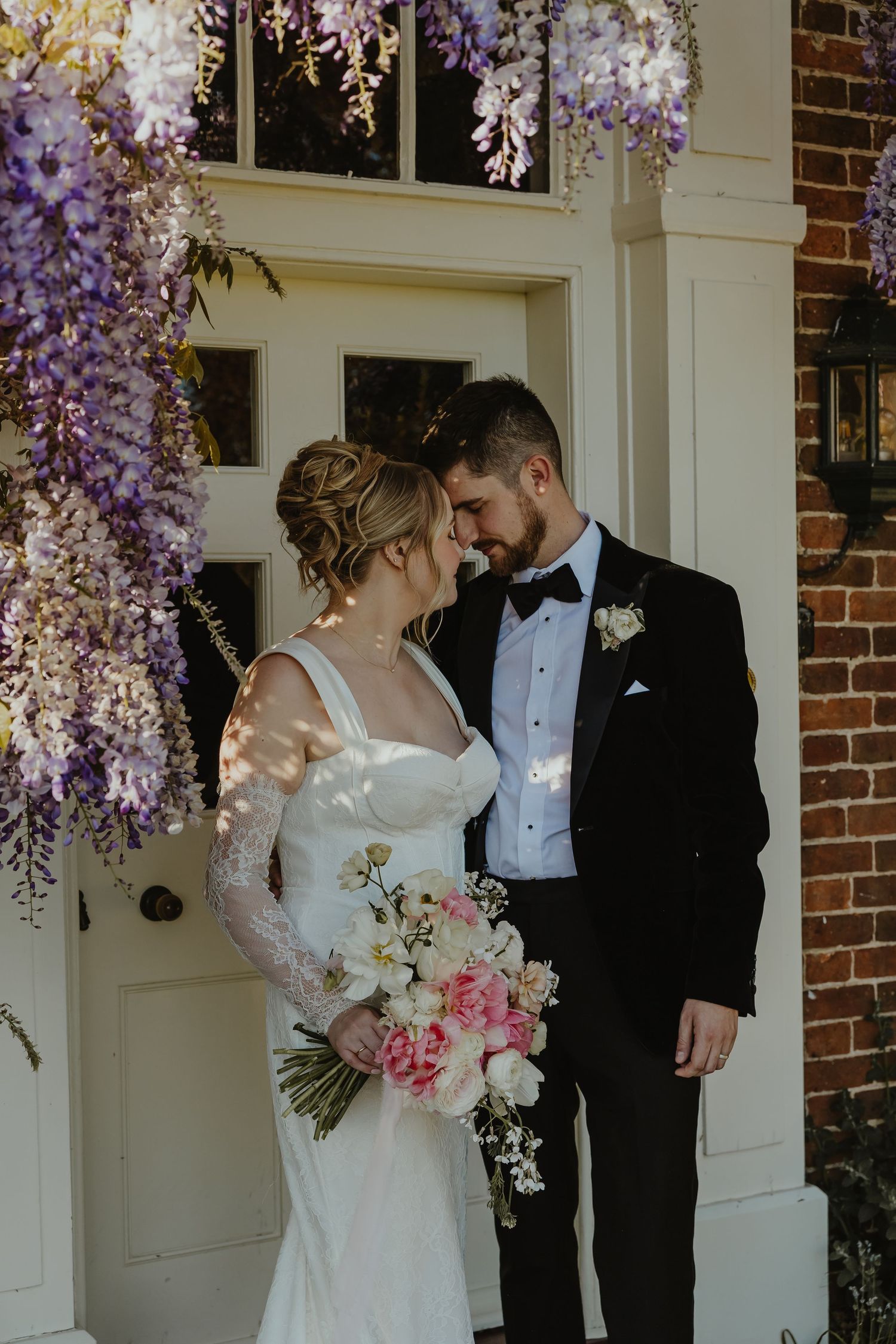 Bride and groom share a romantic moment under blooming wisteria at a white door, she holds a pink and white bouquet.