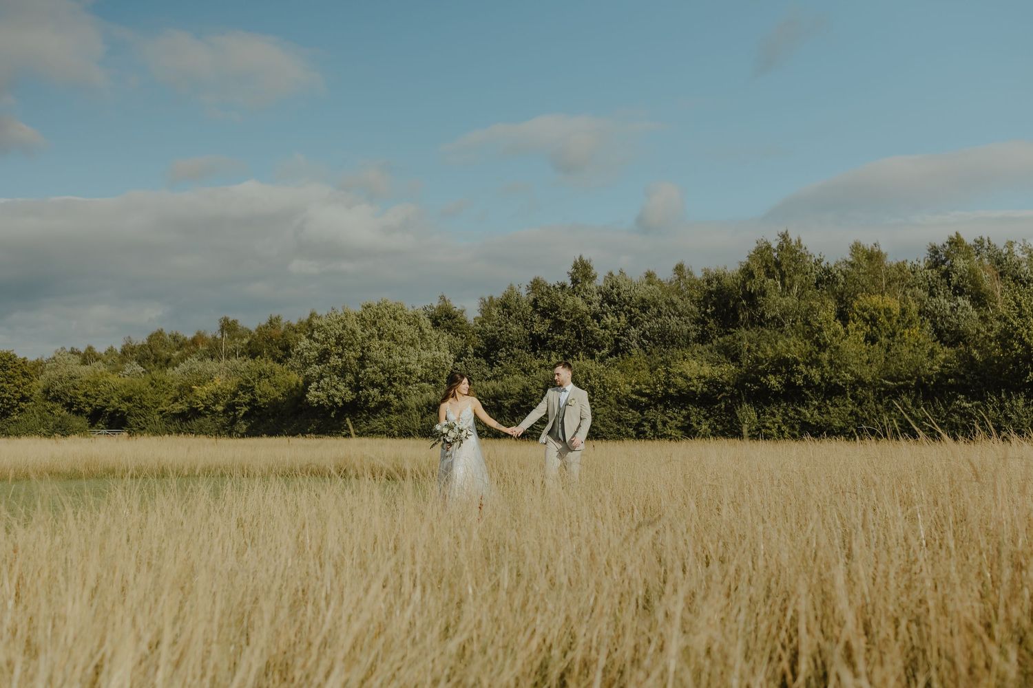 Romantic wedding couple holds hands while walking through tall grass field.