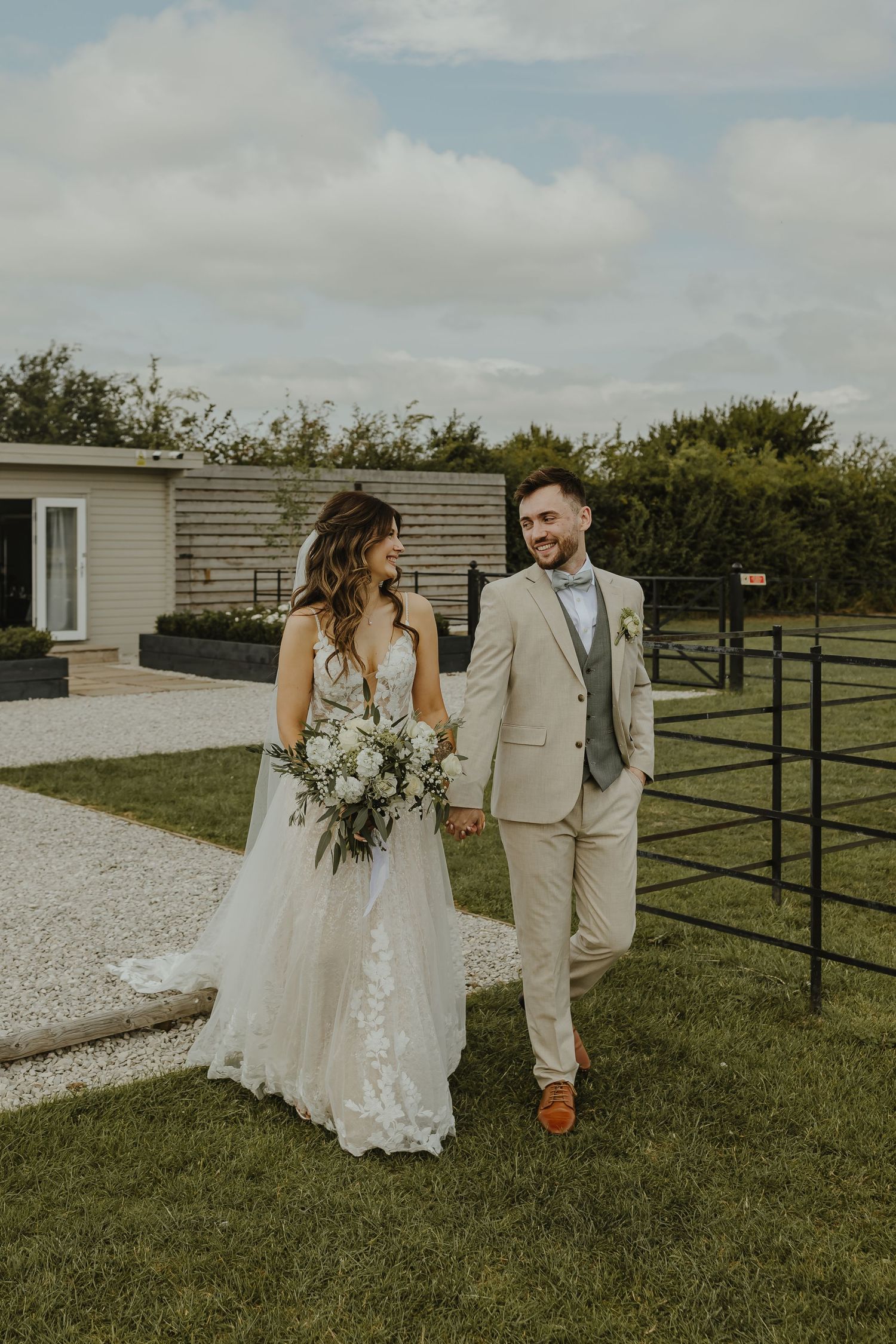 Couple in wedding attire share a candid moment on a gravel path by a white building.