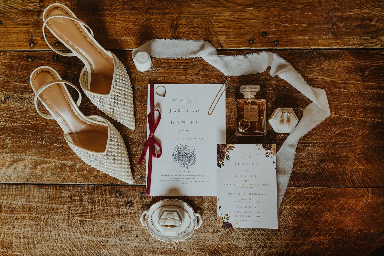 Wedding details flatlay with white shoes, invitation, perfume bottle and ribbons on wooden surface.