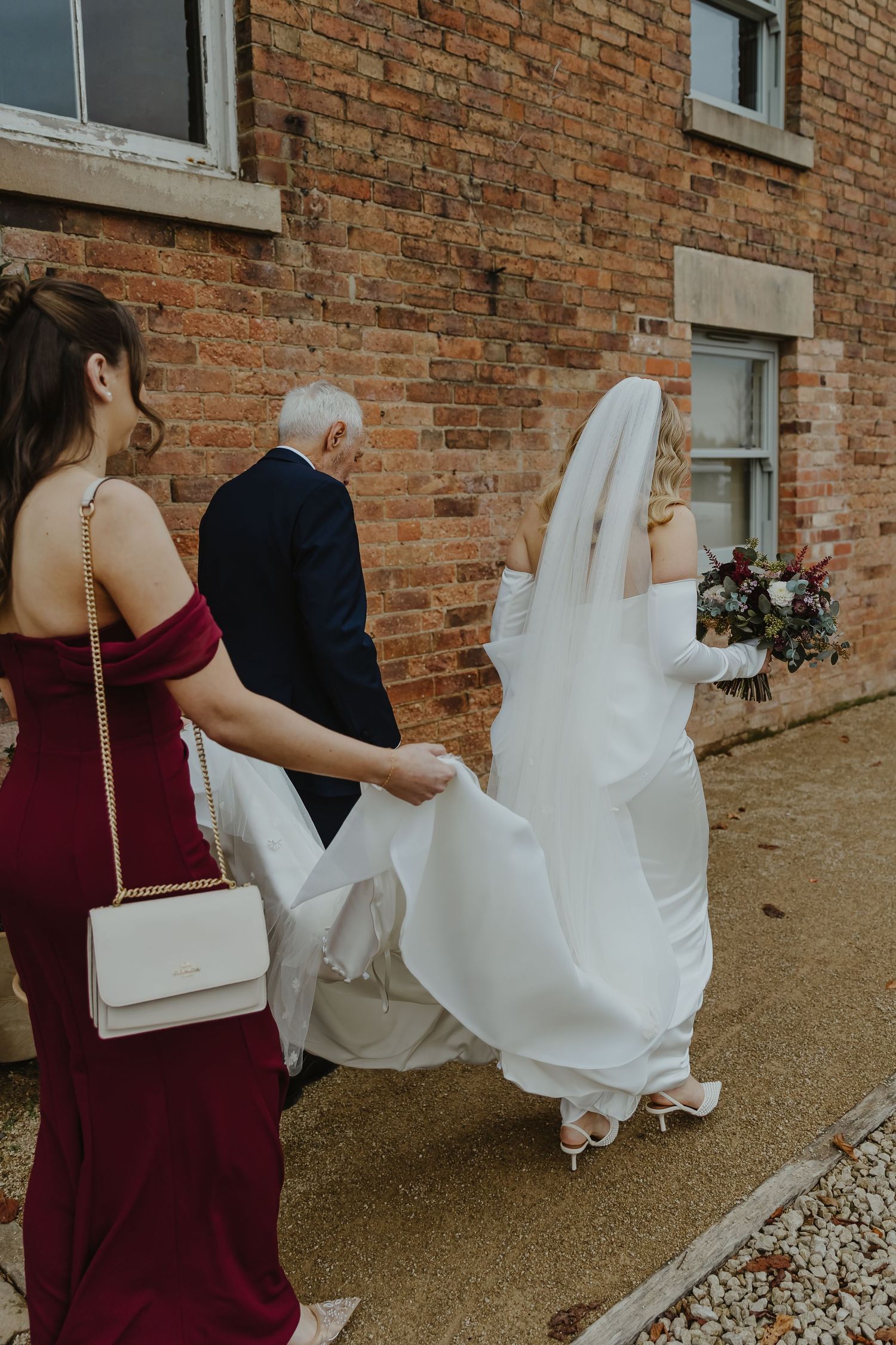 Wedding party walking outside brick building with flowing white dress train.