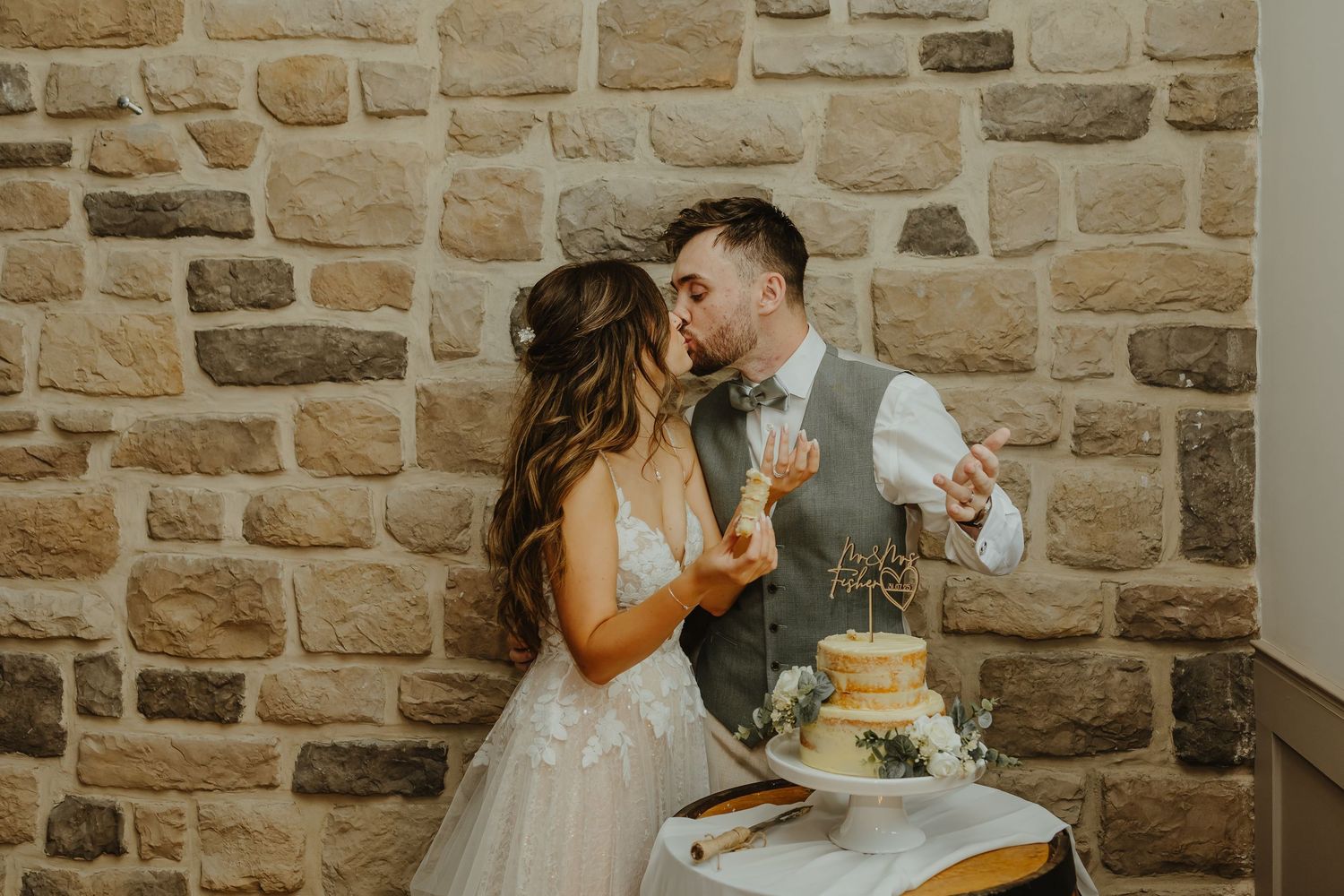 Newlyweds playfully feeding each other wedding cake against a stone wall backdrop.