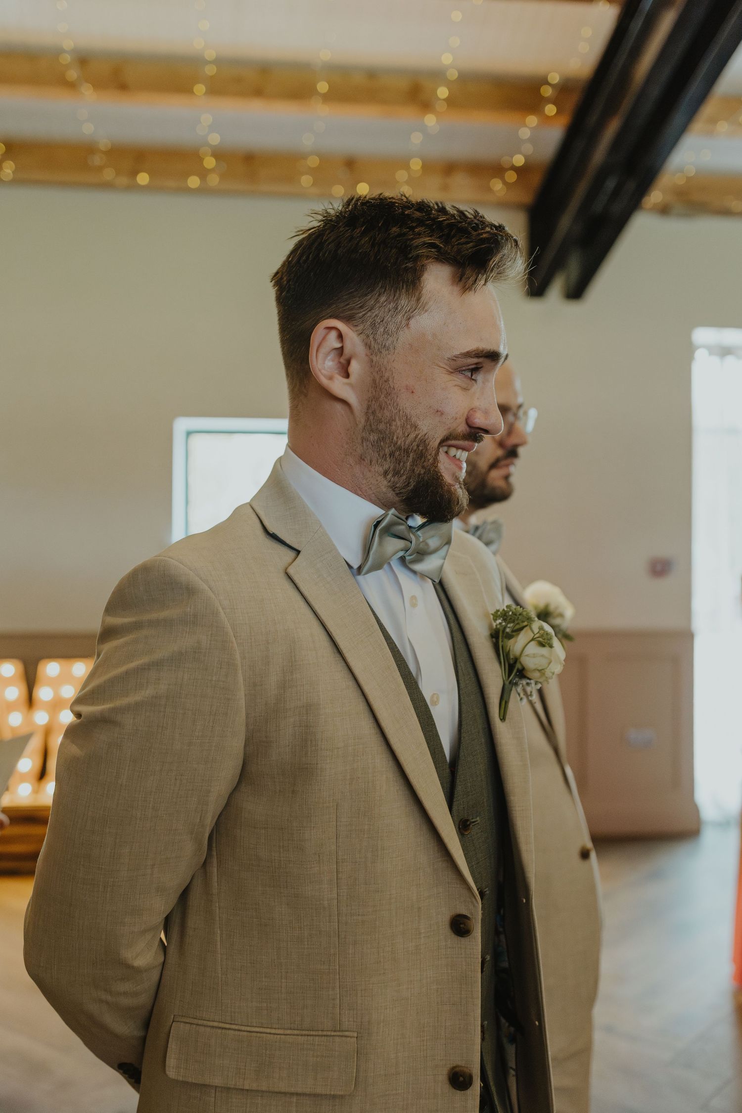 Groomsmen in beige suits and white boutonnières standing in an indoor venue.