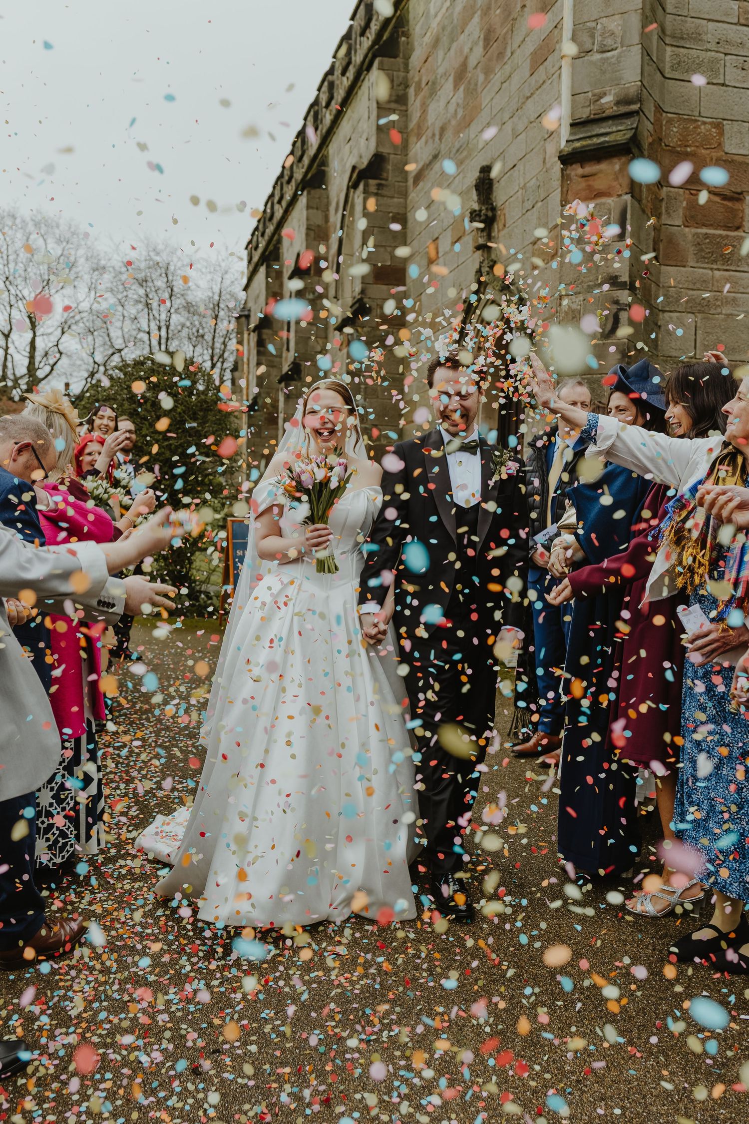Bride and groom walk through colorful confetti shower surrounded by cheering wedding guests outside a stone building.