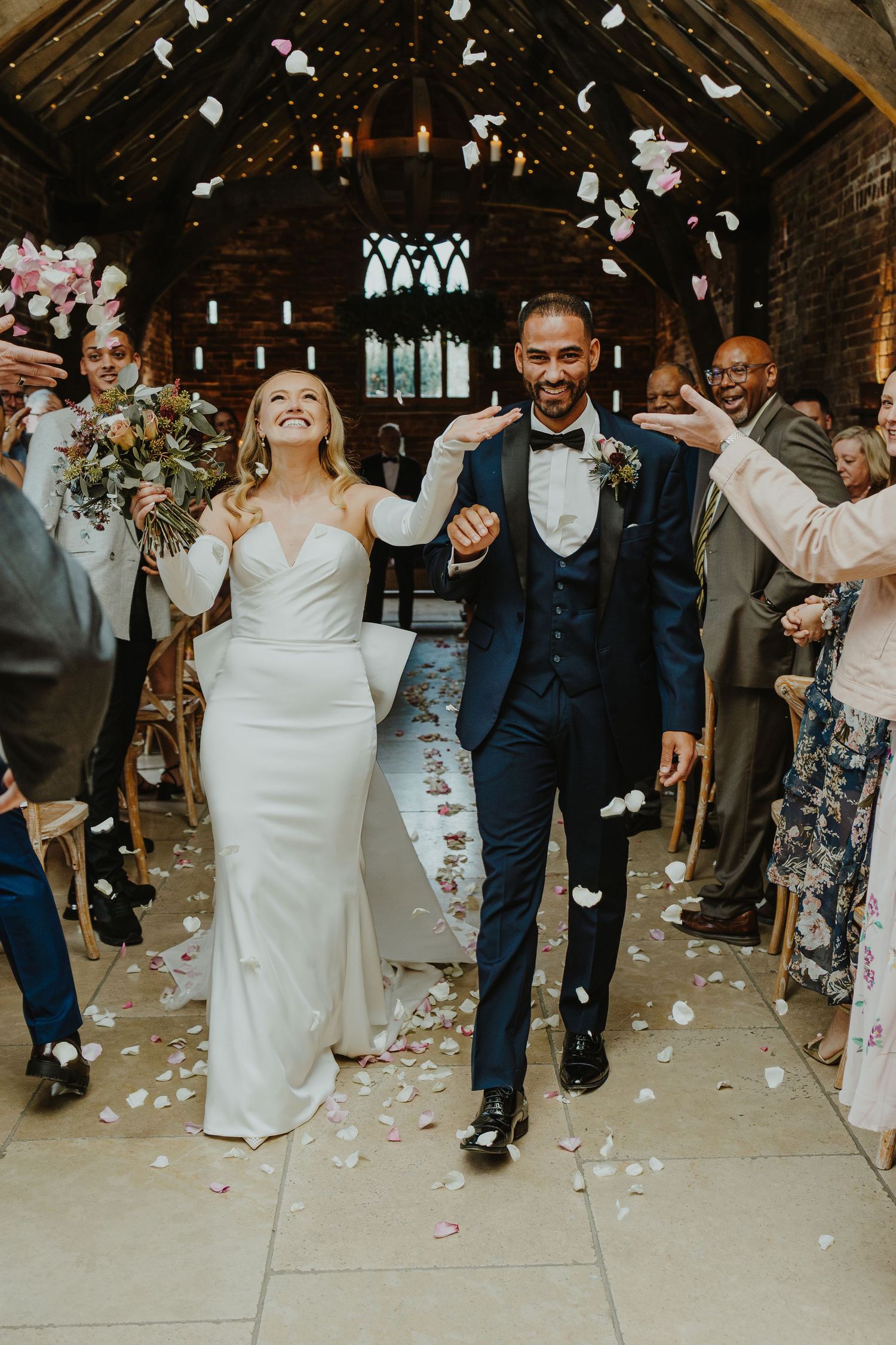 Wedding party walks down church aisle while guests toss flower petals in joyful celebration.