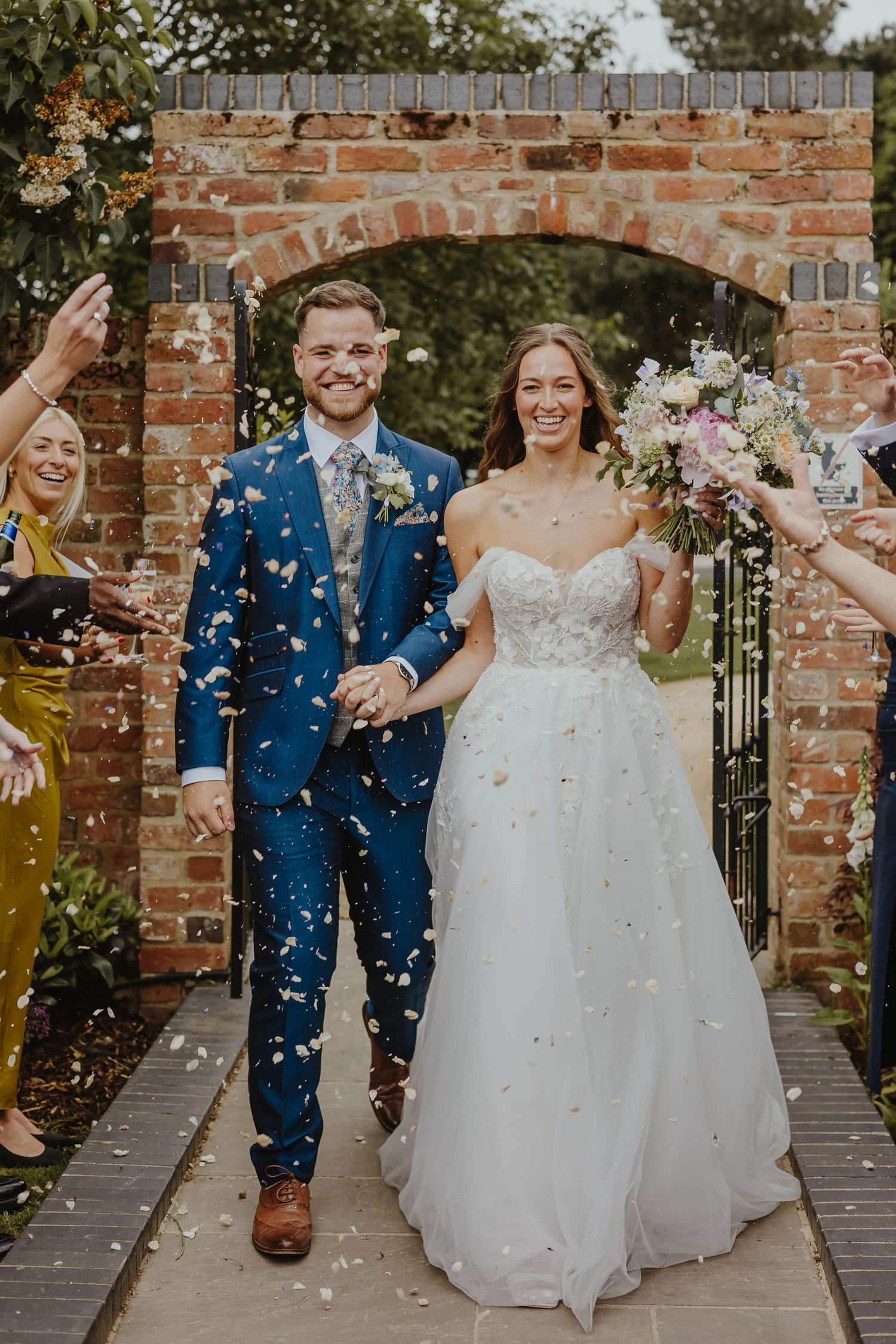 Couple celebrates with confetti toss in front of brick wall at wedding venue.