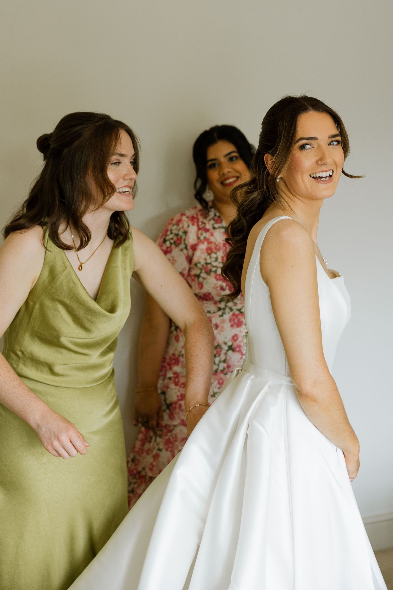 Bride in white gown smiles with bridesmaids in green satin and floral dress during wedding preparations.