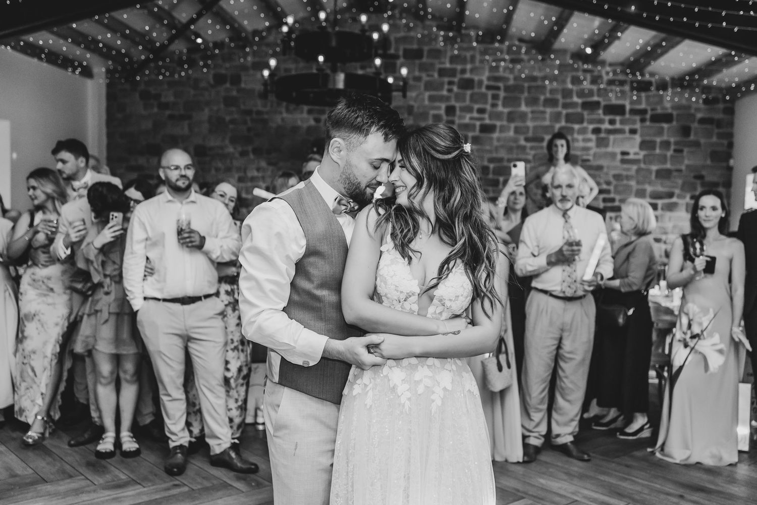 Black and white photo of a couple's first dance surrounded by wedding guests in a rustic venue.