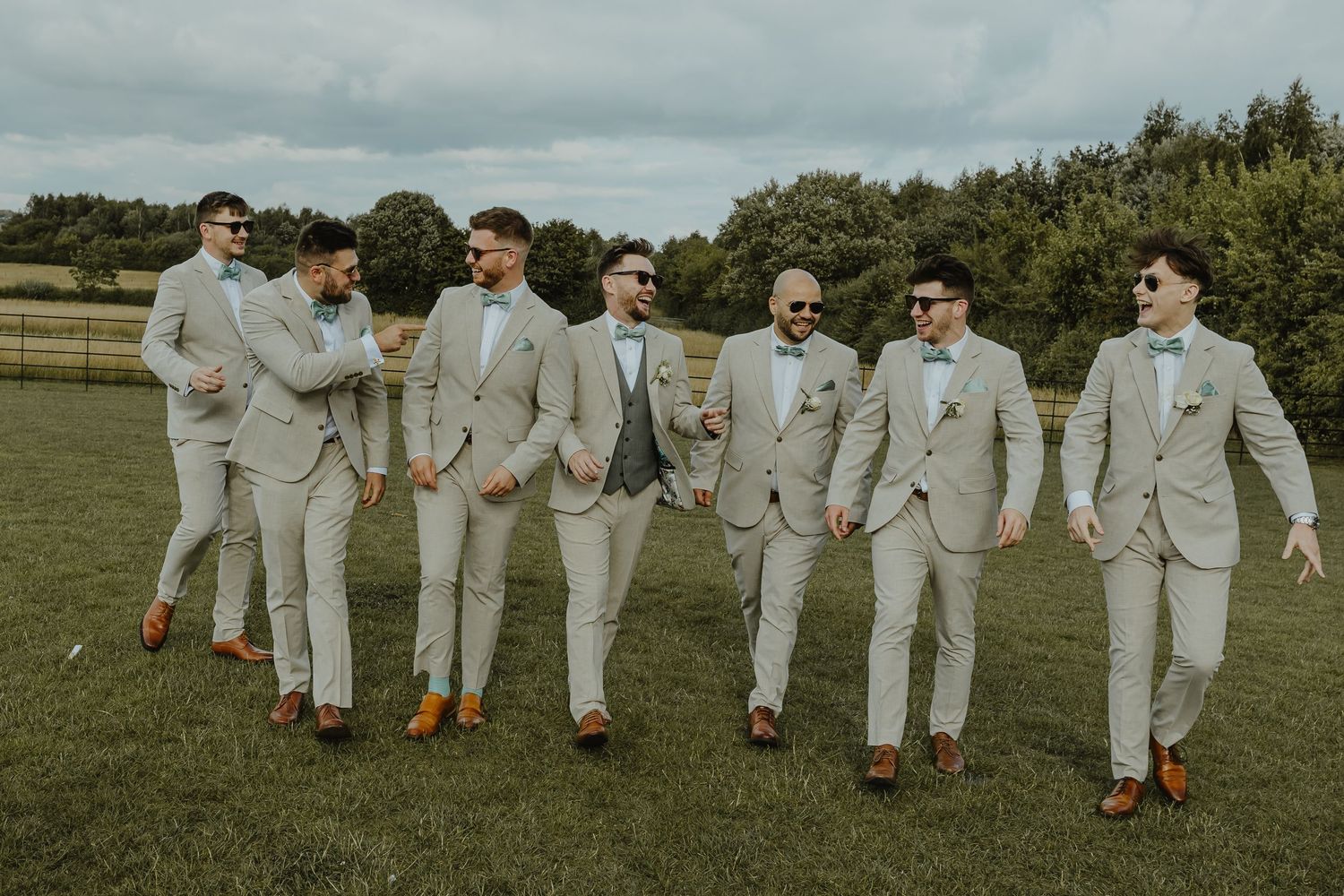 Wedding party groomsmen in light gray suits walking and posing casually on grass field.