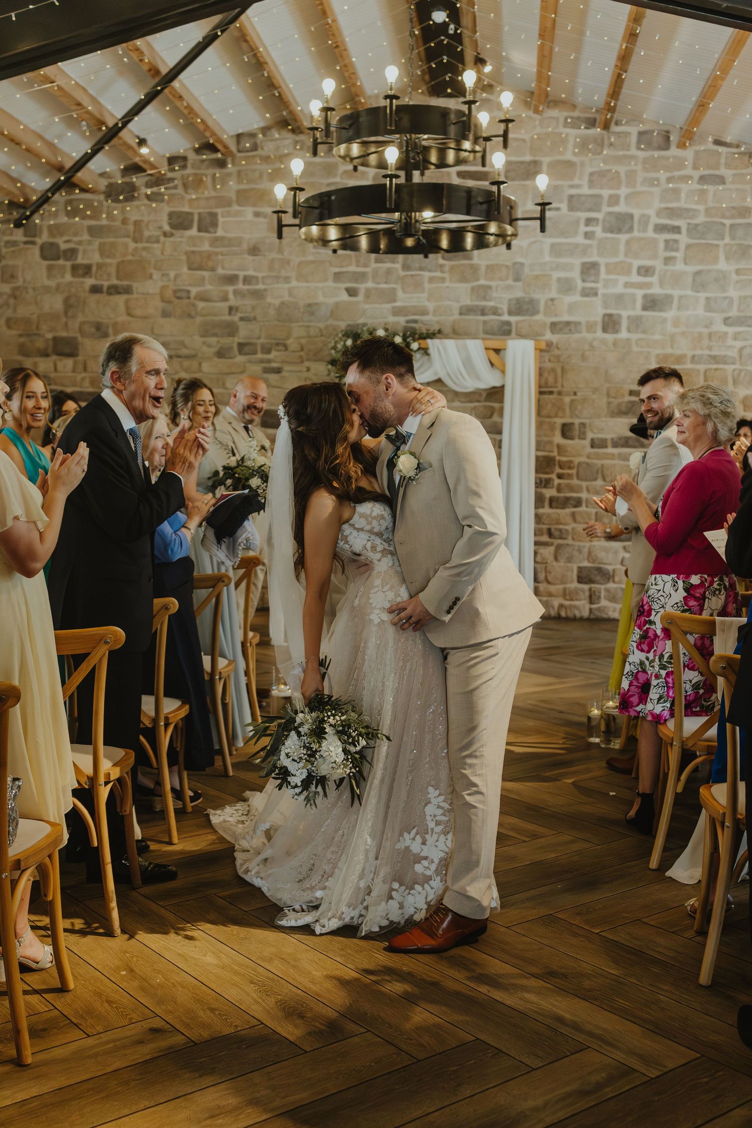 Newlyweds share an intimate moment as guests celebrate in a rustic brick-walled venue.