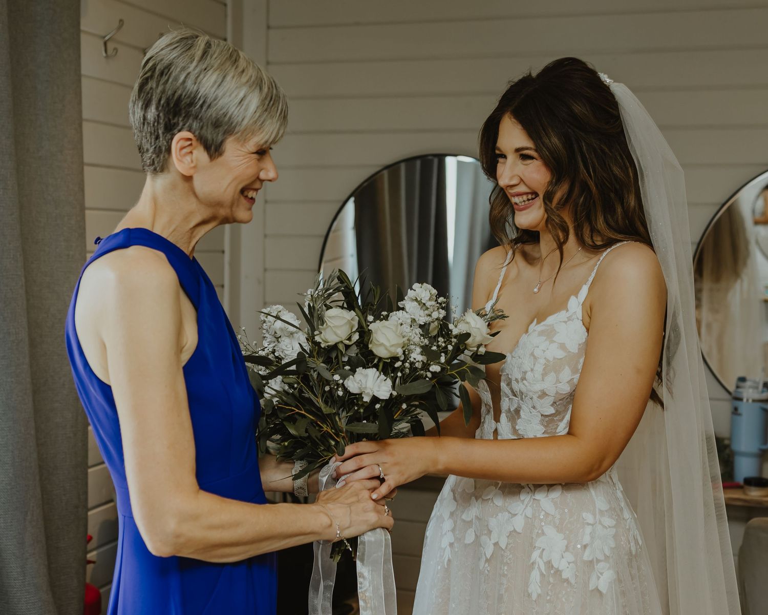 A bride in a lace wedding gown holds a white and green floral bouquet while getting ready.