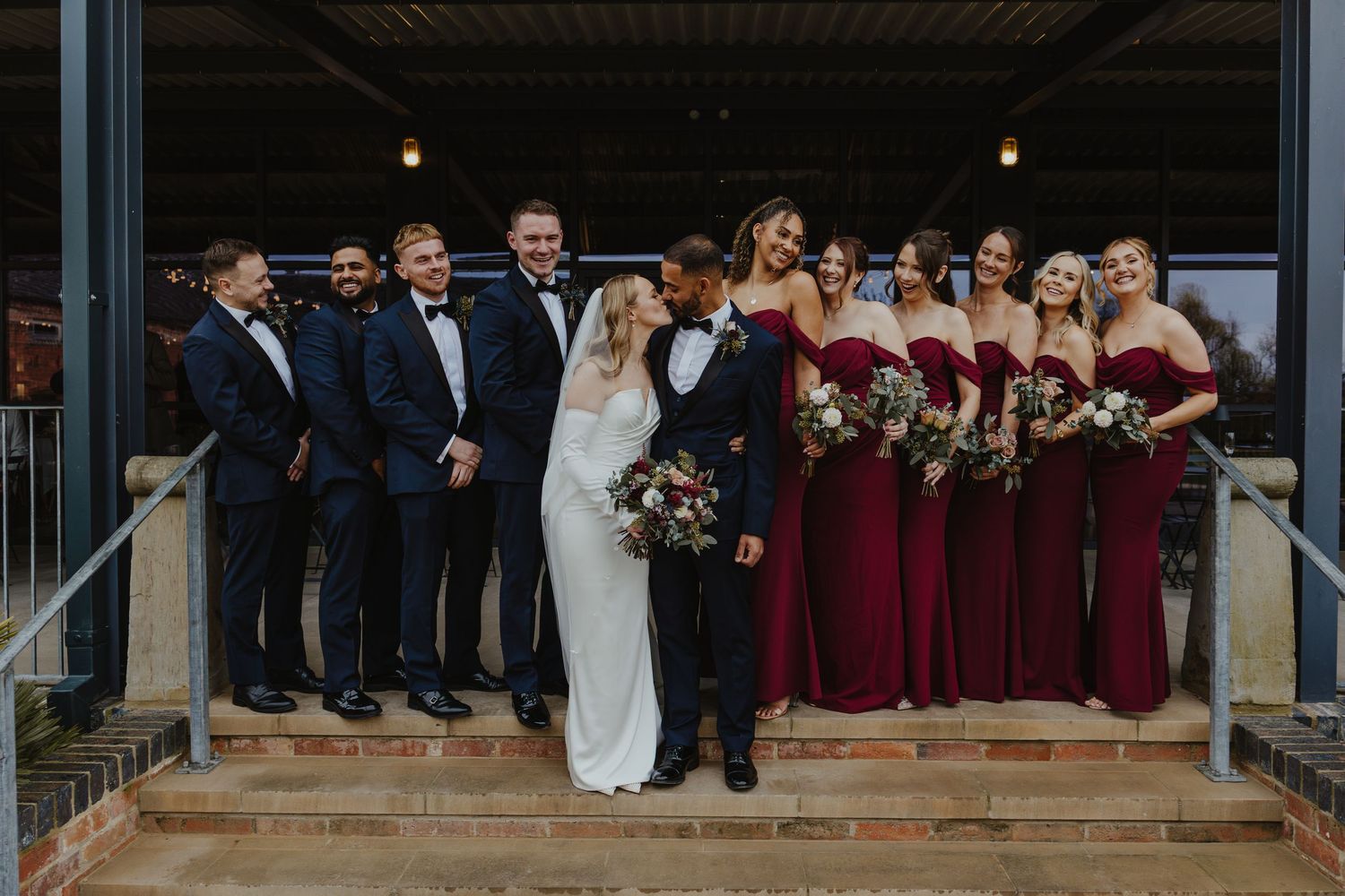 Large wedding party poses on stone steps outside brick building in formal attire.