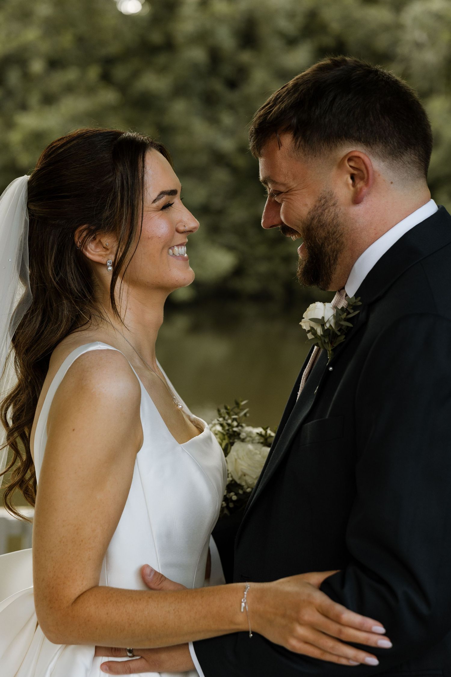 A smiling bride in a white gown and groom in a dark suit share a tender moment outdoors near a pond.