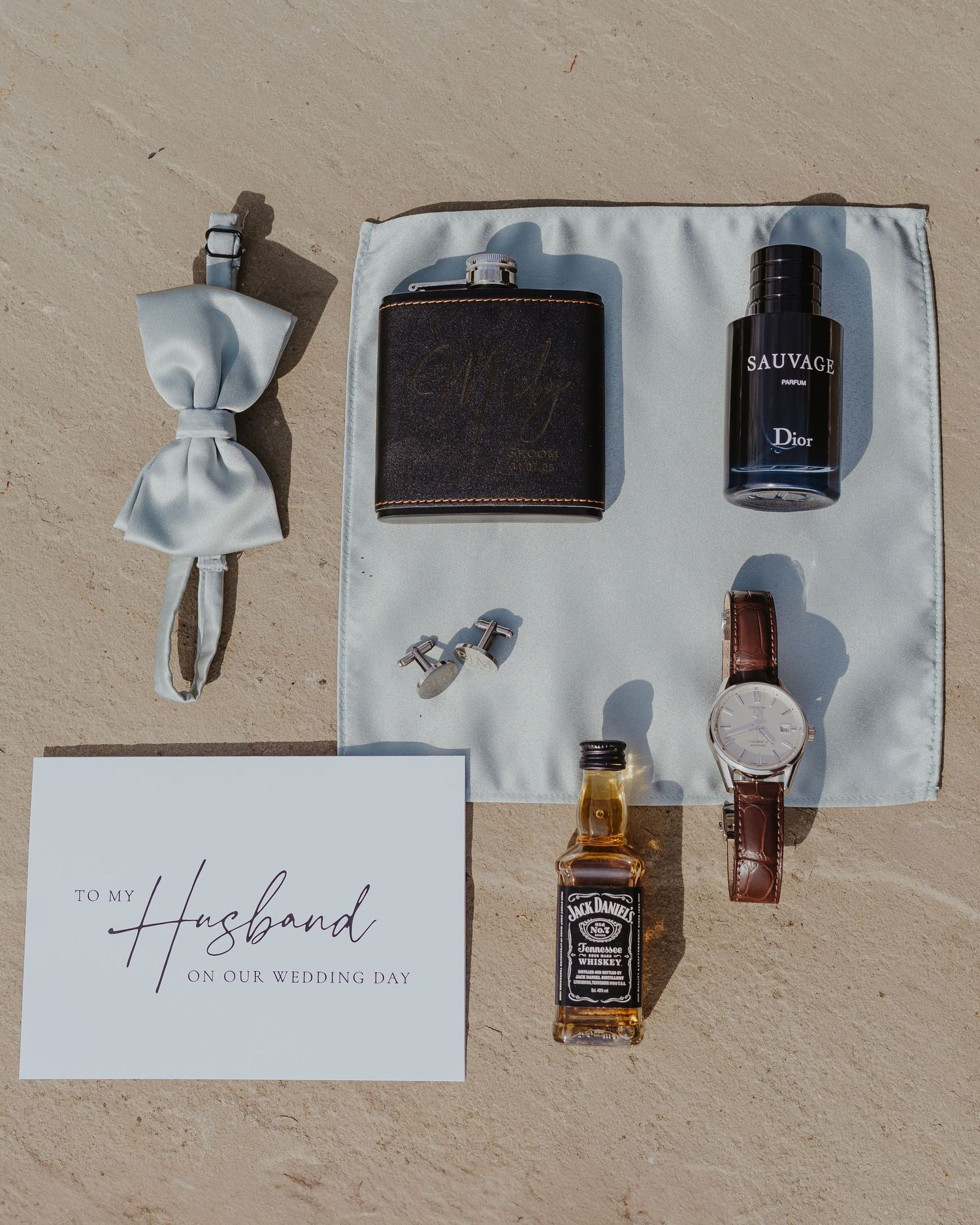 Flat lay of groom's accessories with Hosanna text, leather goods, and a bottle on beige background.