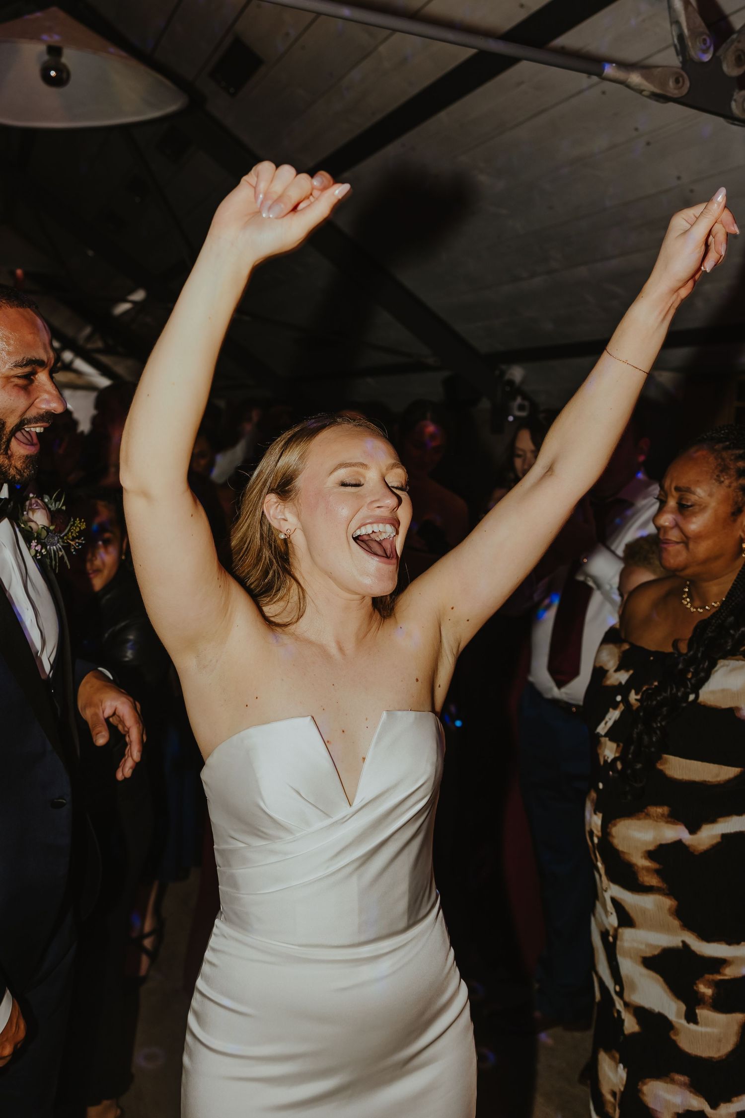 Wedding guests dancing and celebrating with arms raised on a dark dance floor.