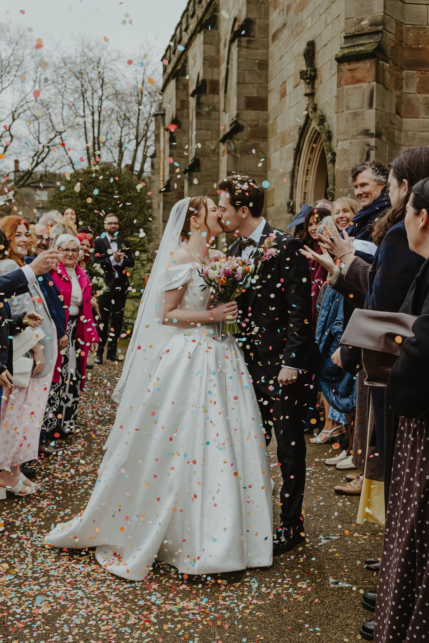 Bride and groom kissing while colorful confetti falls, surrounded by cheering guests outside a stone church.