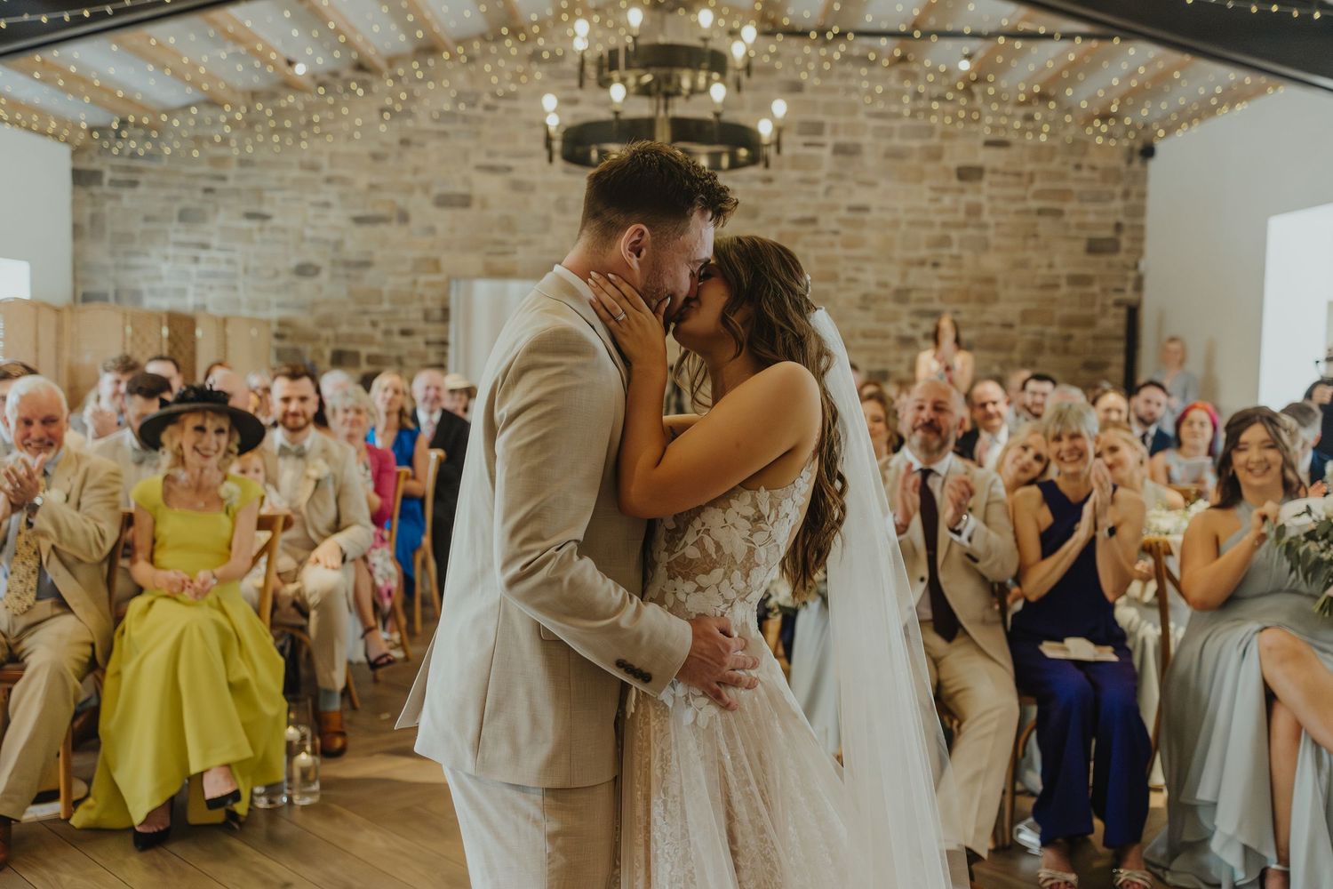 Newlyweds share their first kiss as guests applaud in a rustic venue with stone walls.
