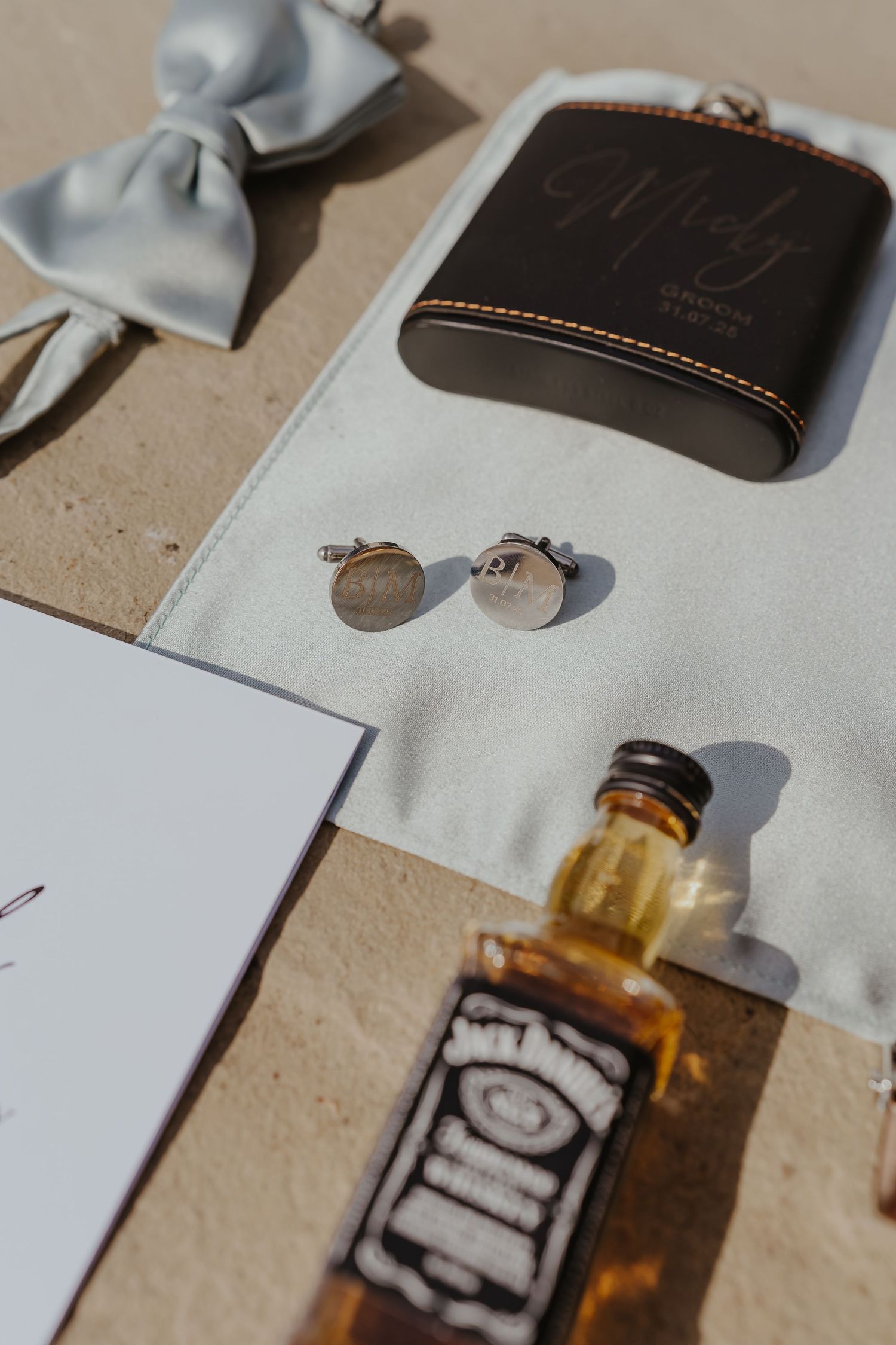 Wedding accessories including a bottle of whiskey, leather wallet, and blue bow tie arranged on a light surface.