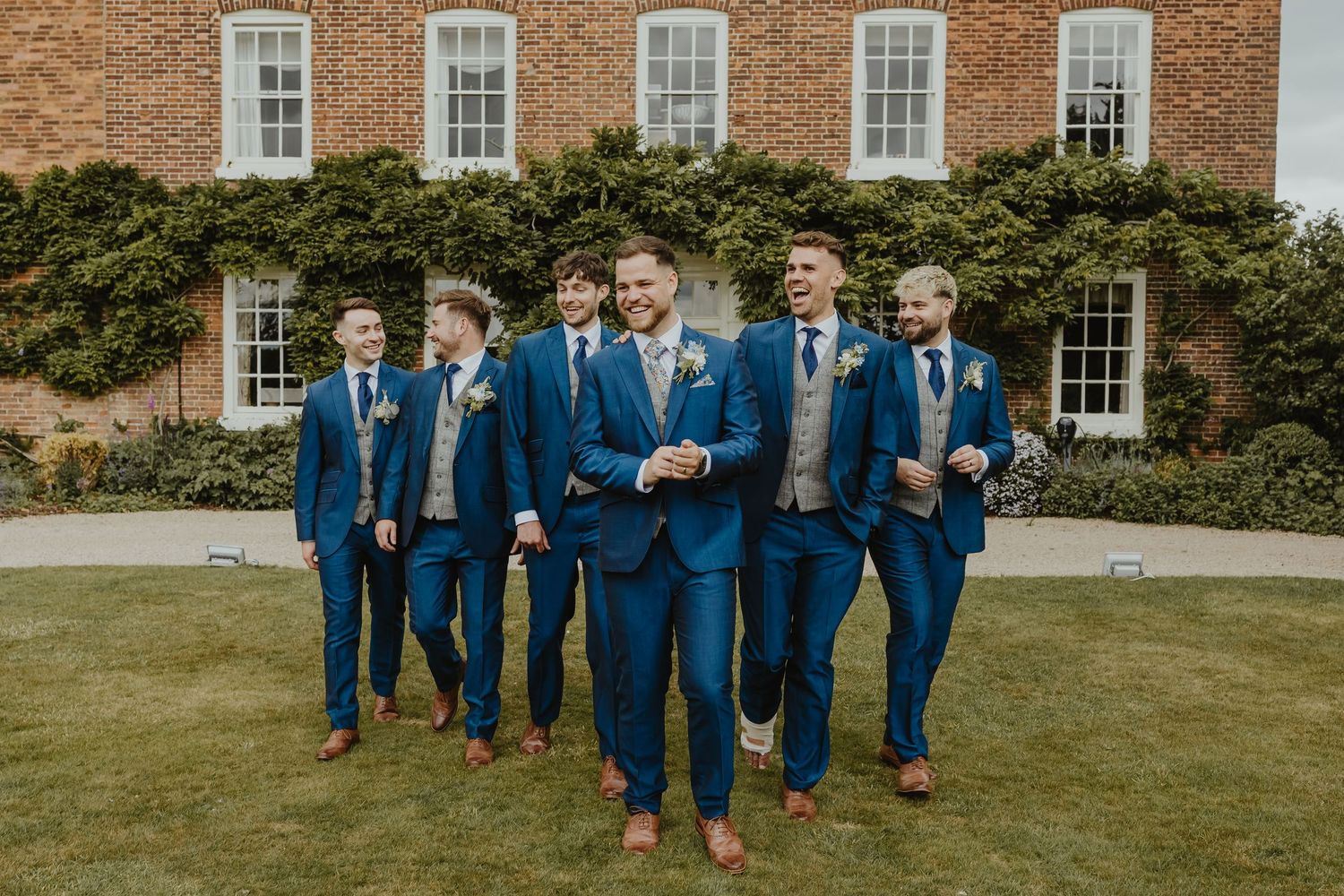 Groomsmen in navy blue suits posing together on a lawn in front of a brick building.