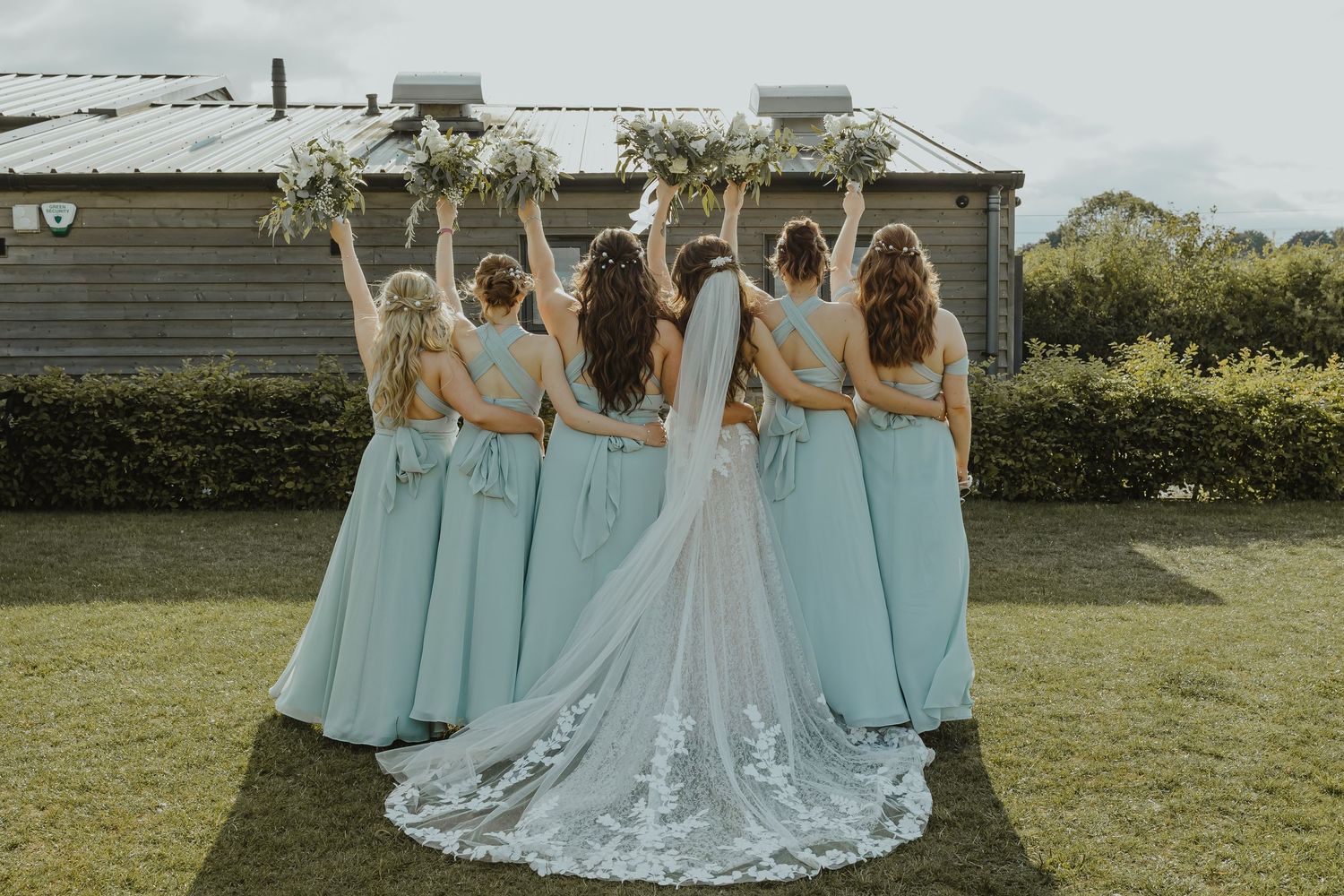 Bridesmaids in light blue dresses pose behind bride showcasing her lace wedding gown train.