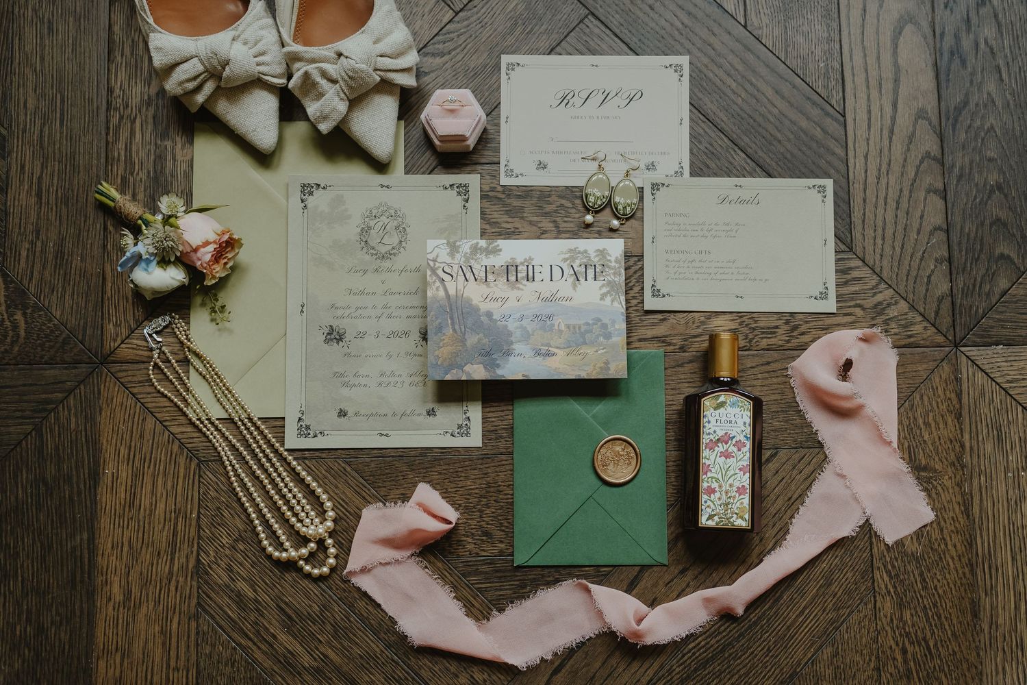Elegant wedding flat lay with green envelopes, wax seal, floral invitations, pearl necklace, pink ribbon, and bridal shoes on wood floor.