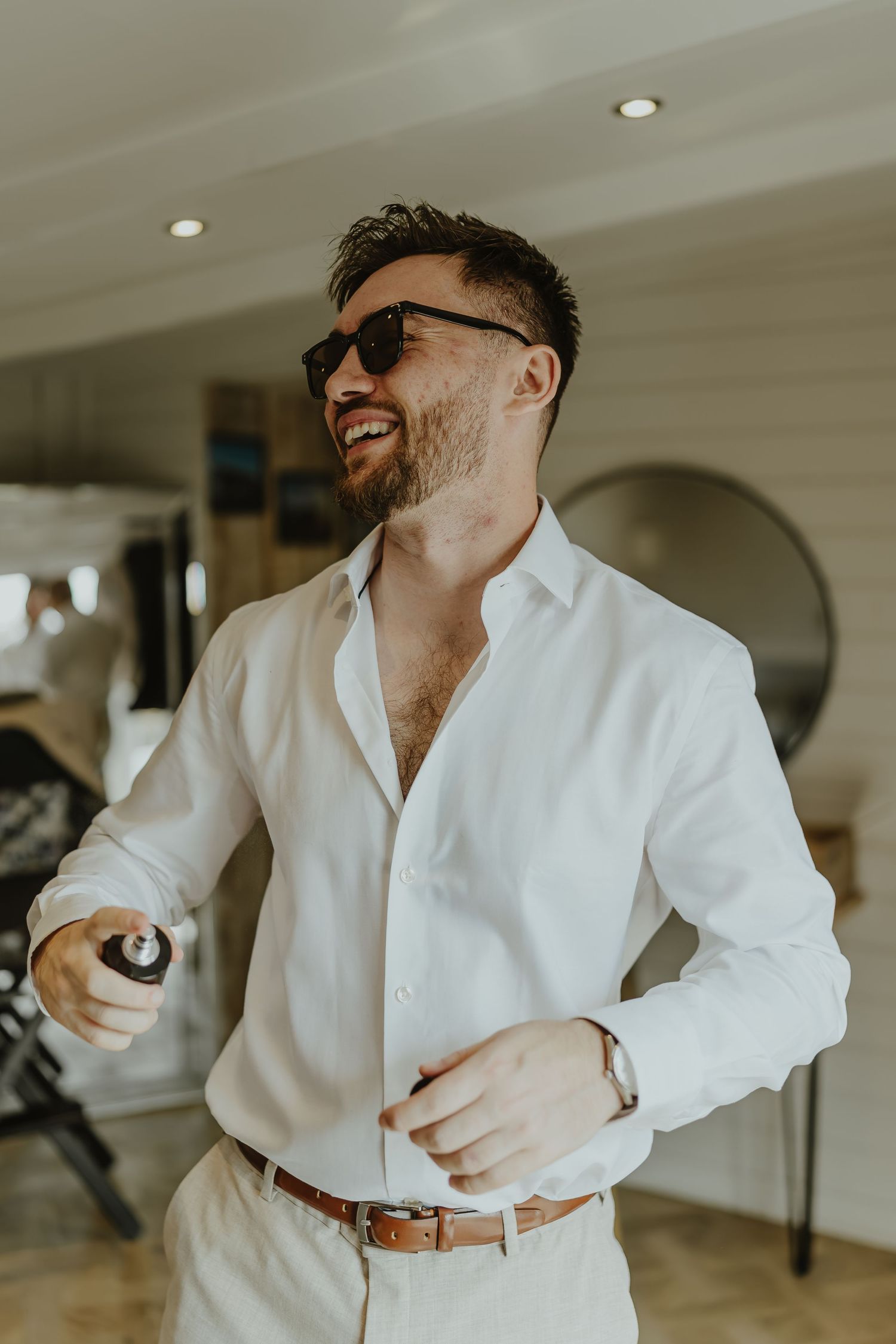 A person in a crisp white shirt and sunglasses laughs while getting dressed for a formal occasion.