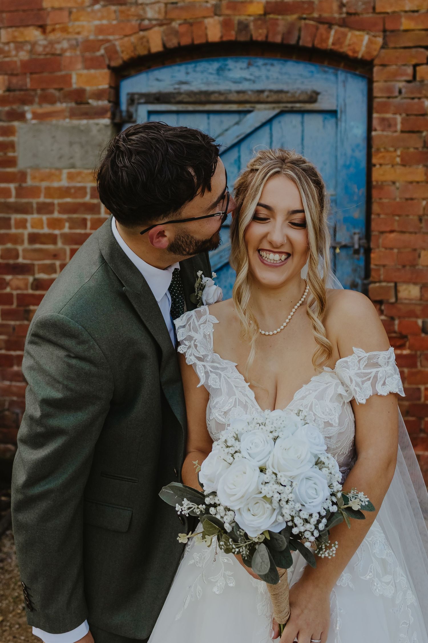 Wedding couple poses against rustic brick wall with blue door backdrop.
