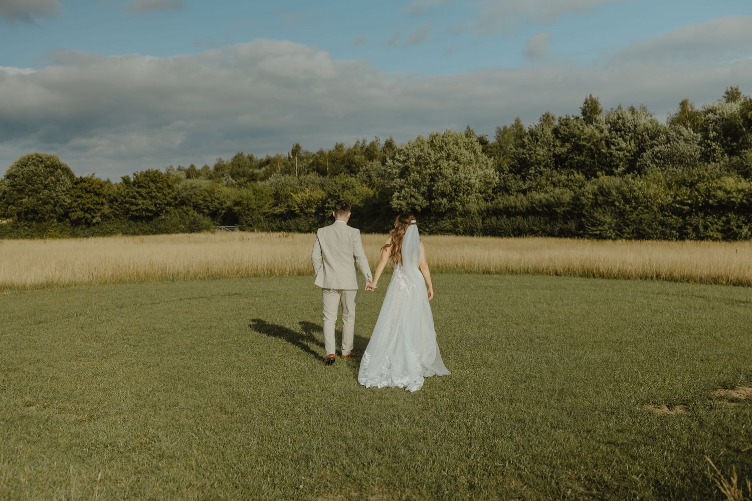 Couple in wedding attire walking hand in hand across a sunlit meadow.