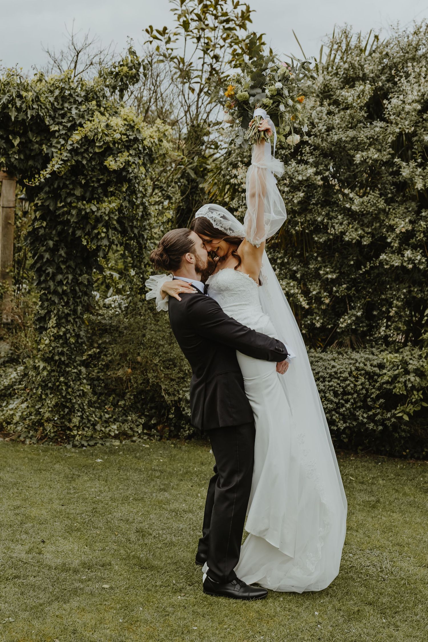 Groom lifts bride in white gown and veil, holding bouquet aloft, sharing a joyful moment in a lush garden setting.