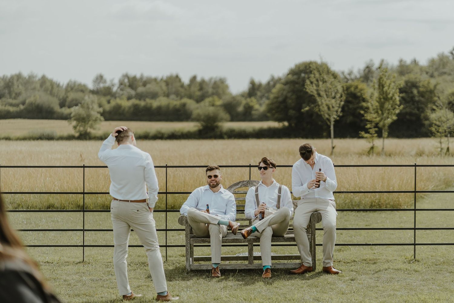 Wedding party relaxing outdoors on benches in an open field with rustic fencing.