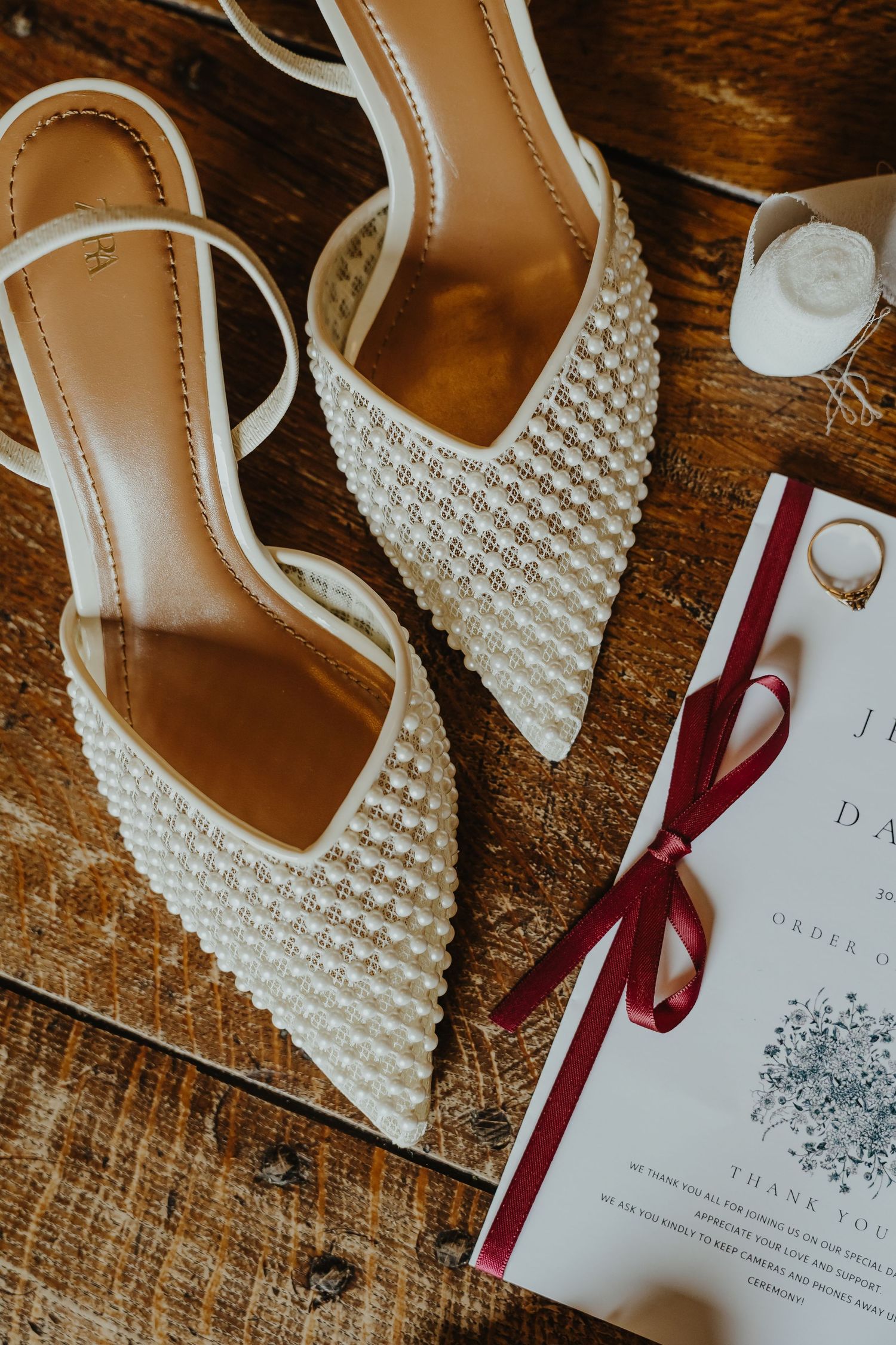 Elegant white bridal shoes with pearl details and wedding invitation with burgundy ribbon on rustic wooden surface.