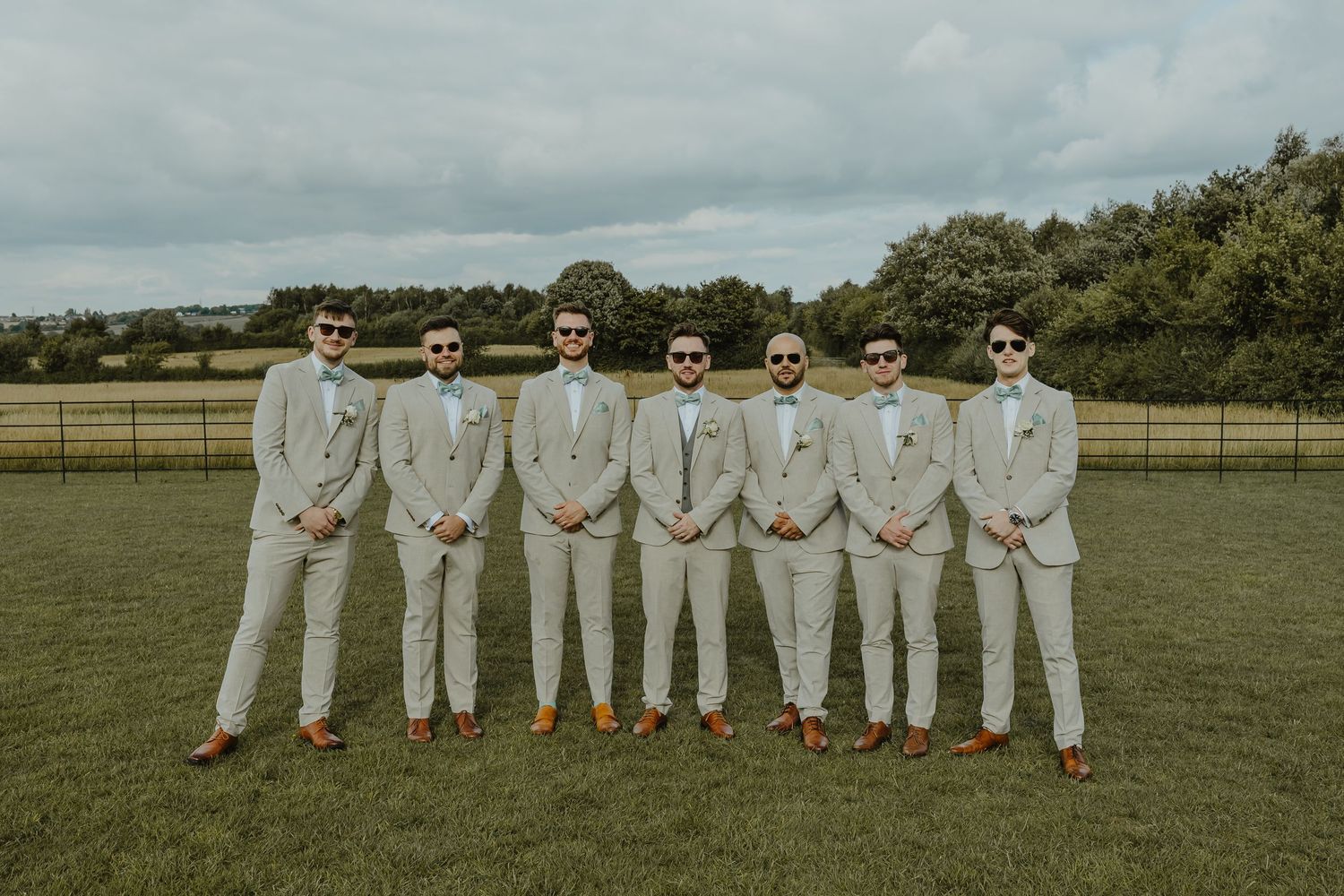 Groomsmen in matching light gray suits and brown shoes stand in line on grassy field.