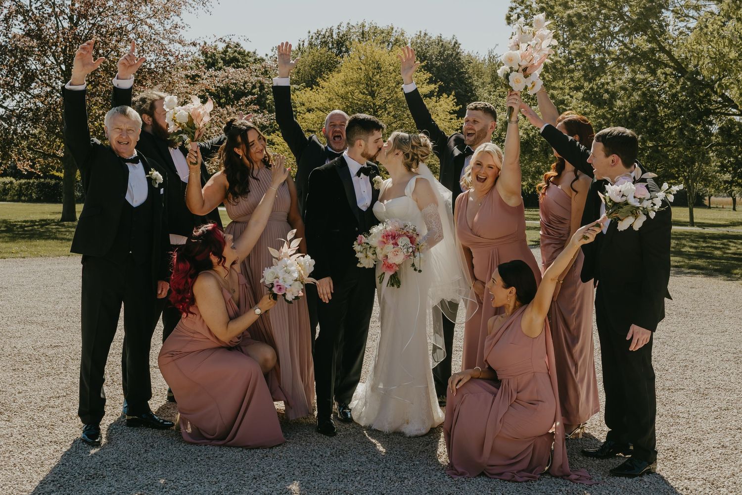 Wedding party celebrating outdoors, bridesmaids in dusty rose gowns, groomsmen in tuxedos, bride and groom kissing while guests cheer.
