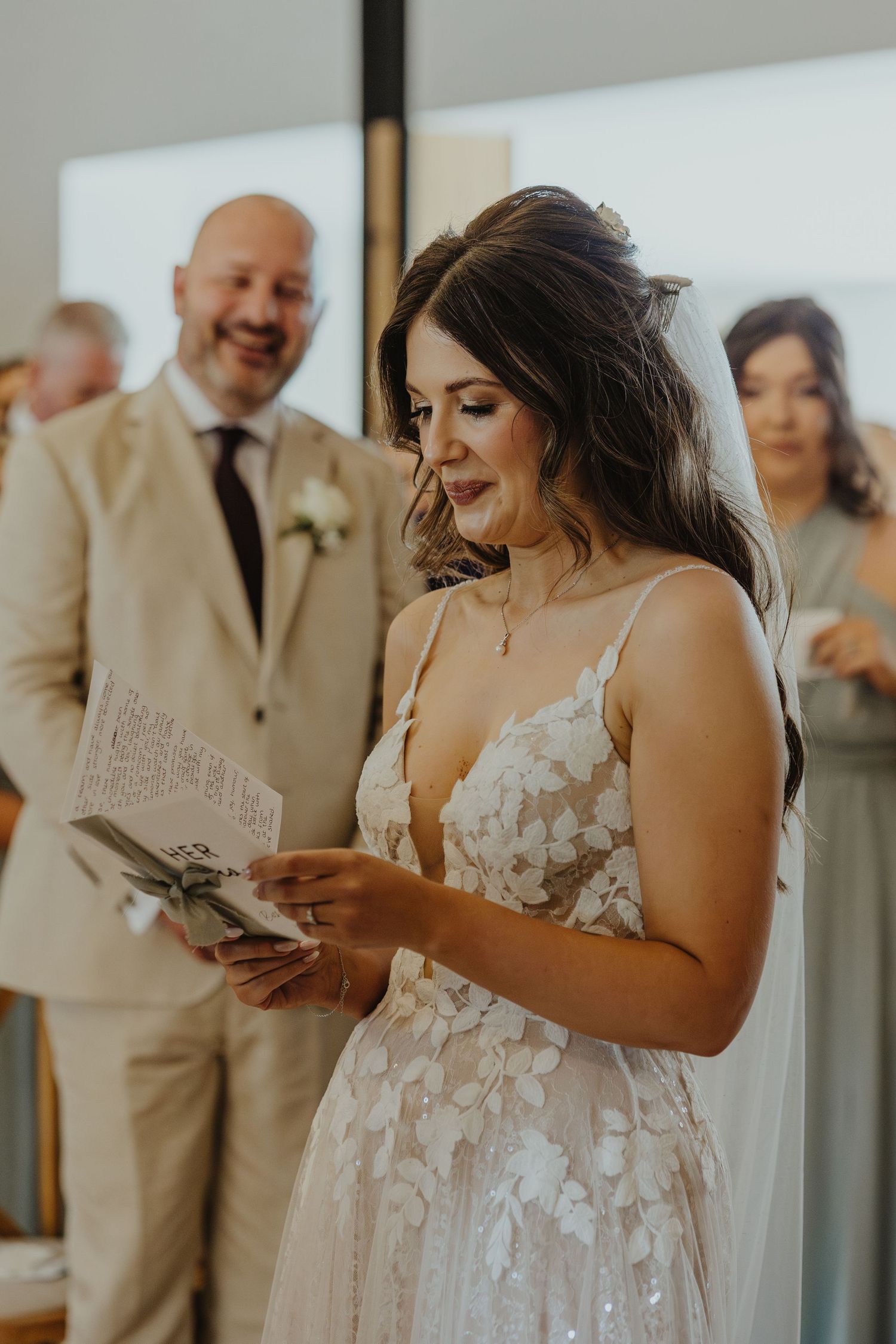 A bride in a floral lace wedding dress reads from a paper during the ceremony.