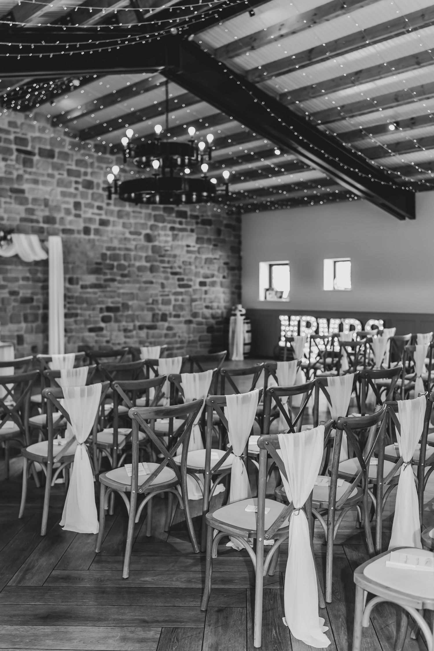 Black and white photo of wedding venue with cross-back chairs and string lights.