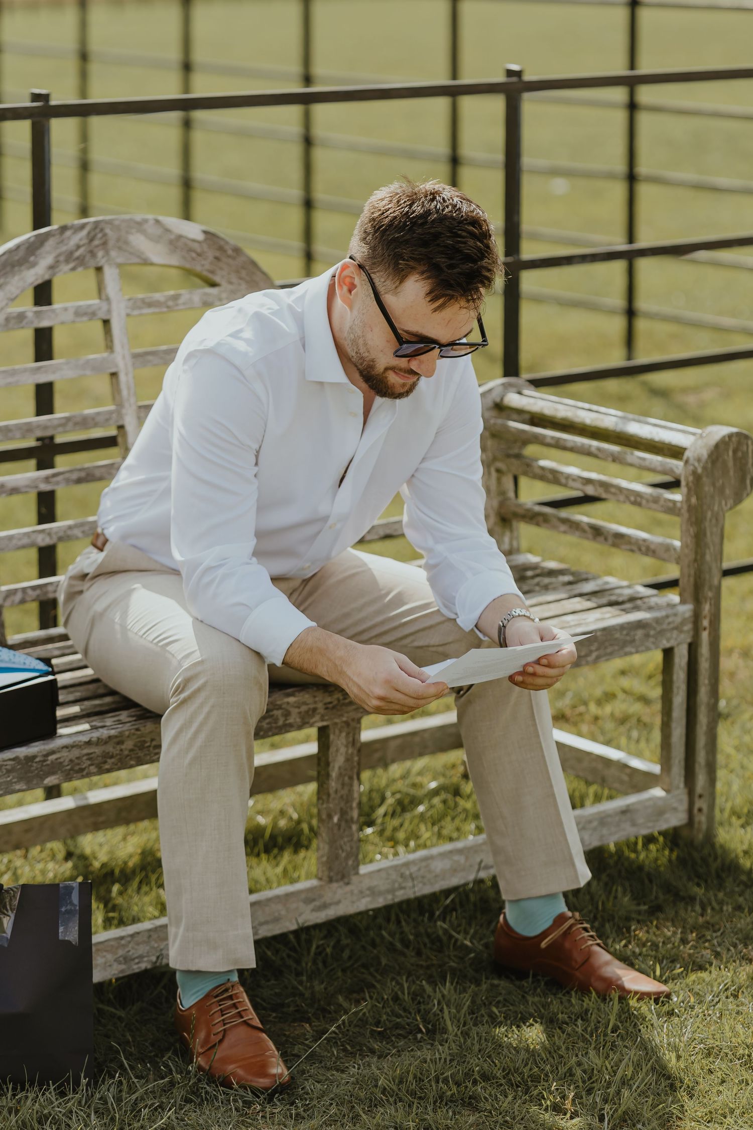 Someone in white shirt and khaki pants sitting on wooden bleachers reading a letter outdoors.