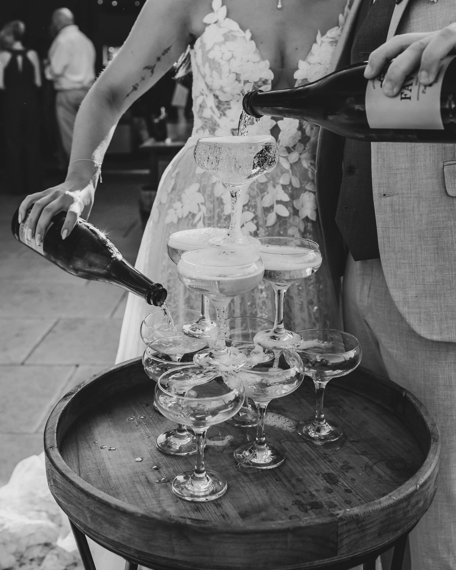 Black and white photo of champagne being poured into vintage coupe glasses on a wooden barrel.