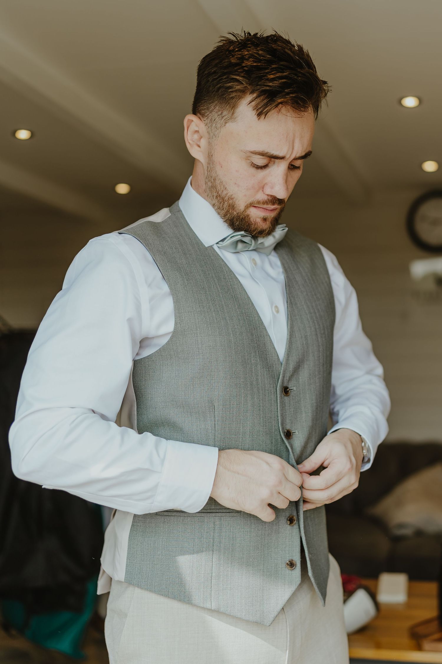 Someone buttons up a light grey vest over a white dress shirt while preparing for a formal event.